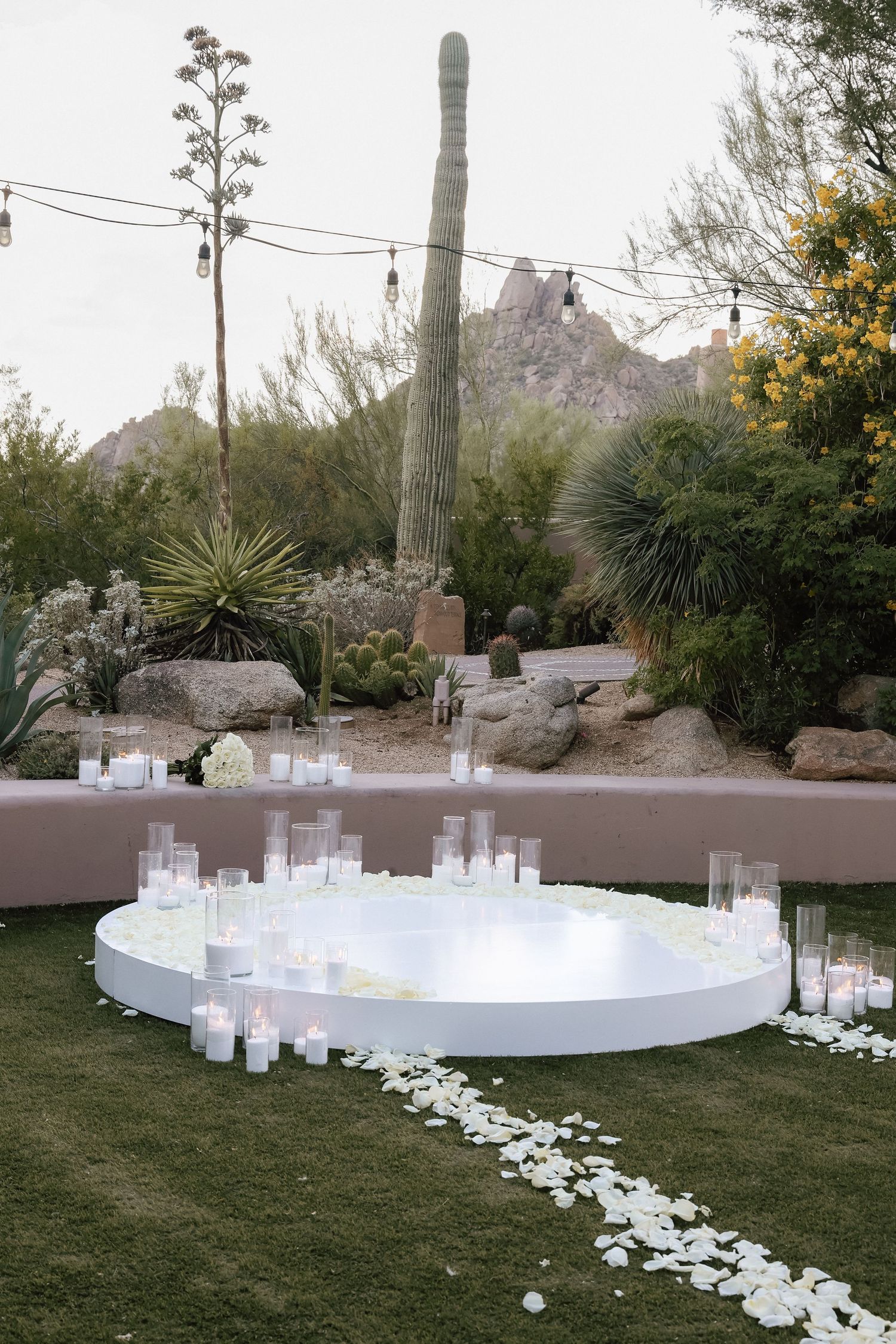 Outdoor desert wedding ceremony setup with a round white platform, white candles, and rose petals surrounded by cacti.