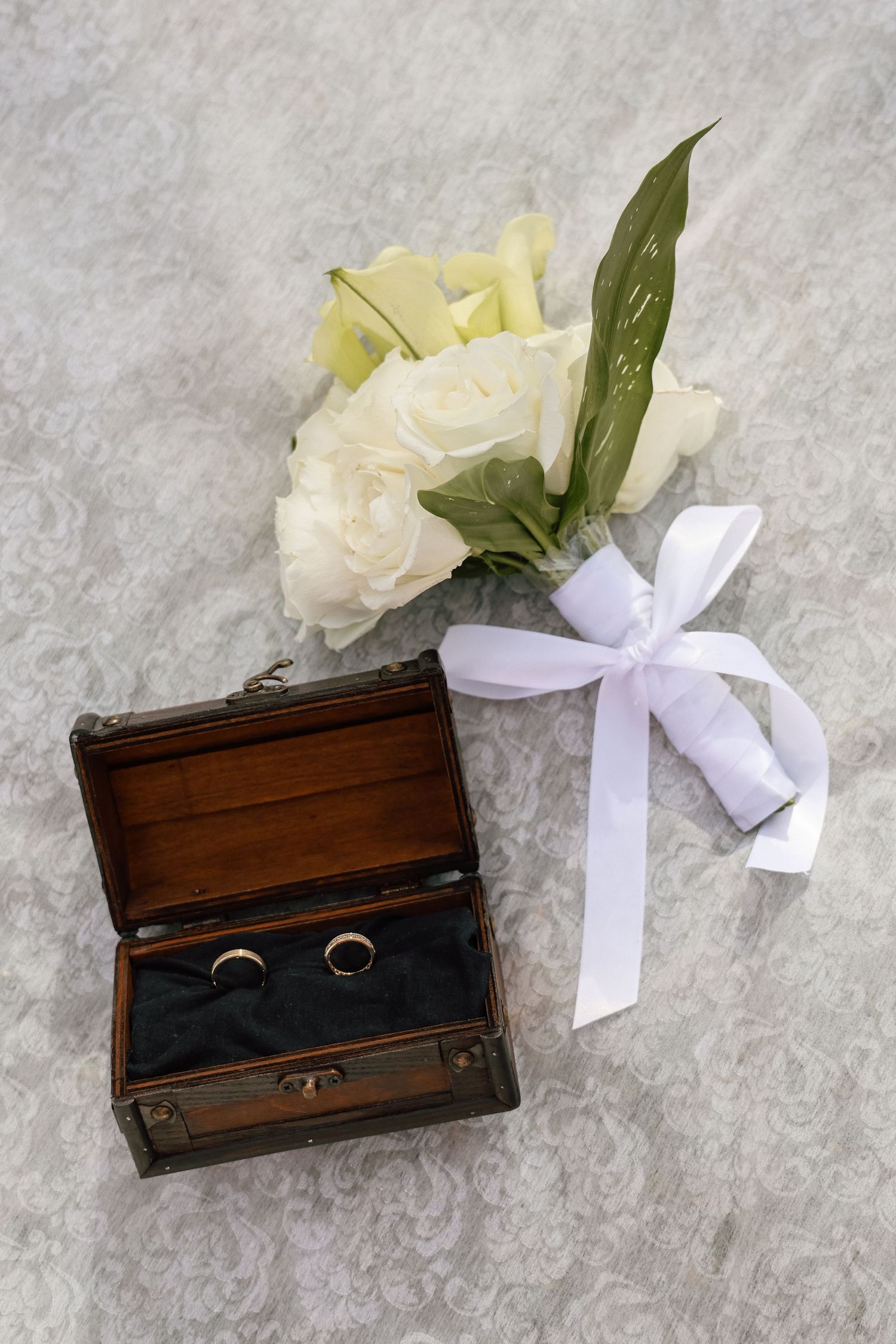 Wedding rings in a wooden chest beside a white rose bouquet tied with a satin ribbon on lace fabric.