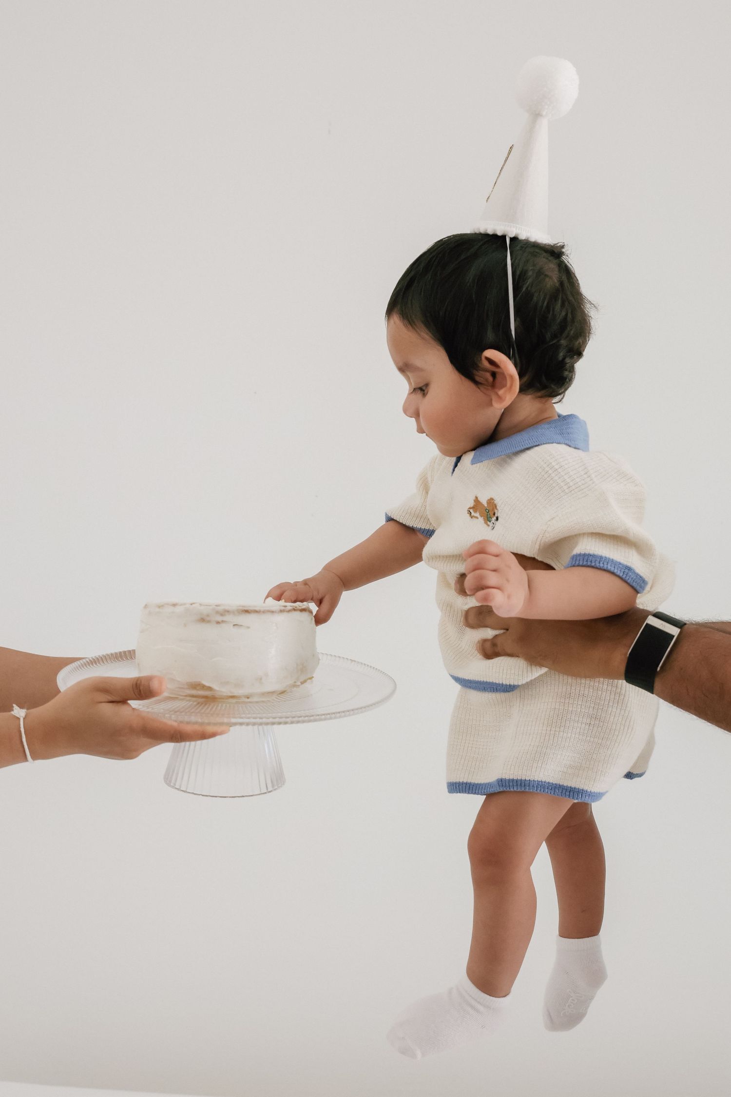 A baby in a party hat and cream outfit reaches for a white cake on a pedestal stand held by an adult.