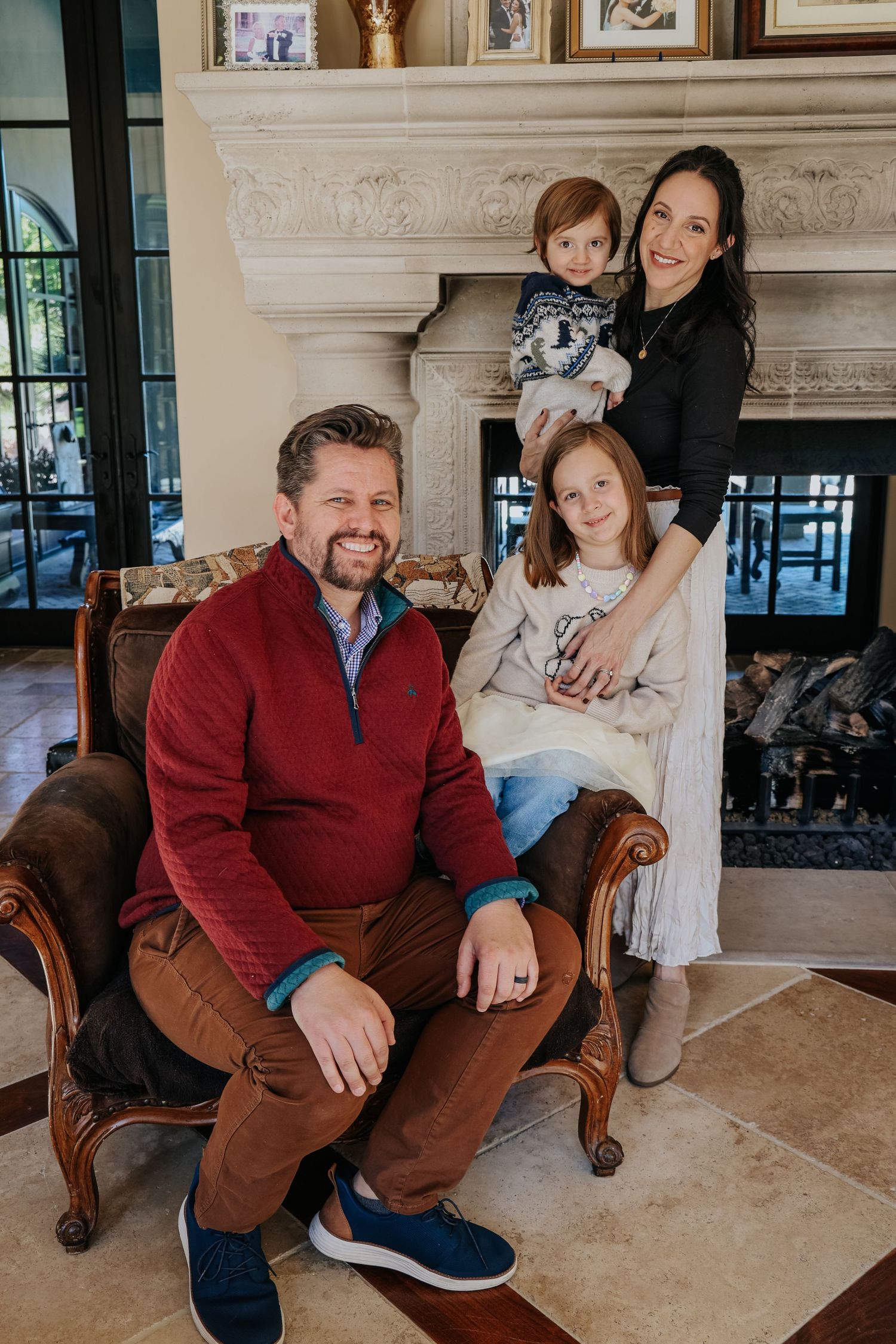 Family of four posing indoors by a fireplace, parents with two children in a cozy living room setting.