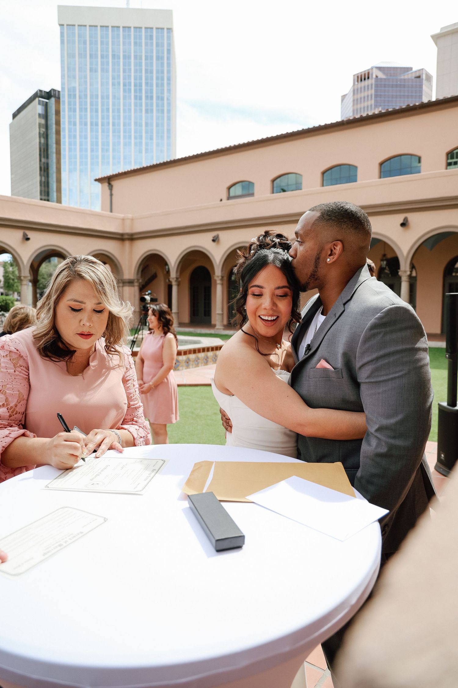 Bride and groom share a kiss while a woman in pink signs wedding documents at an outdoor ceremony venue.