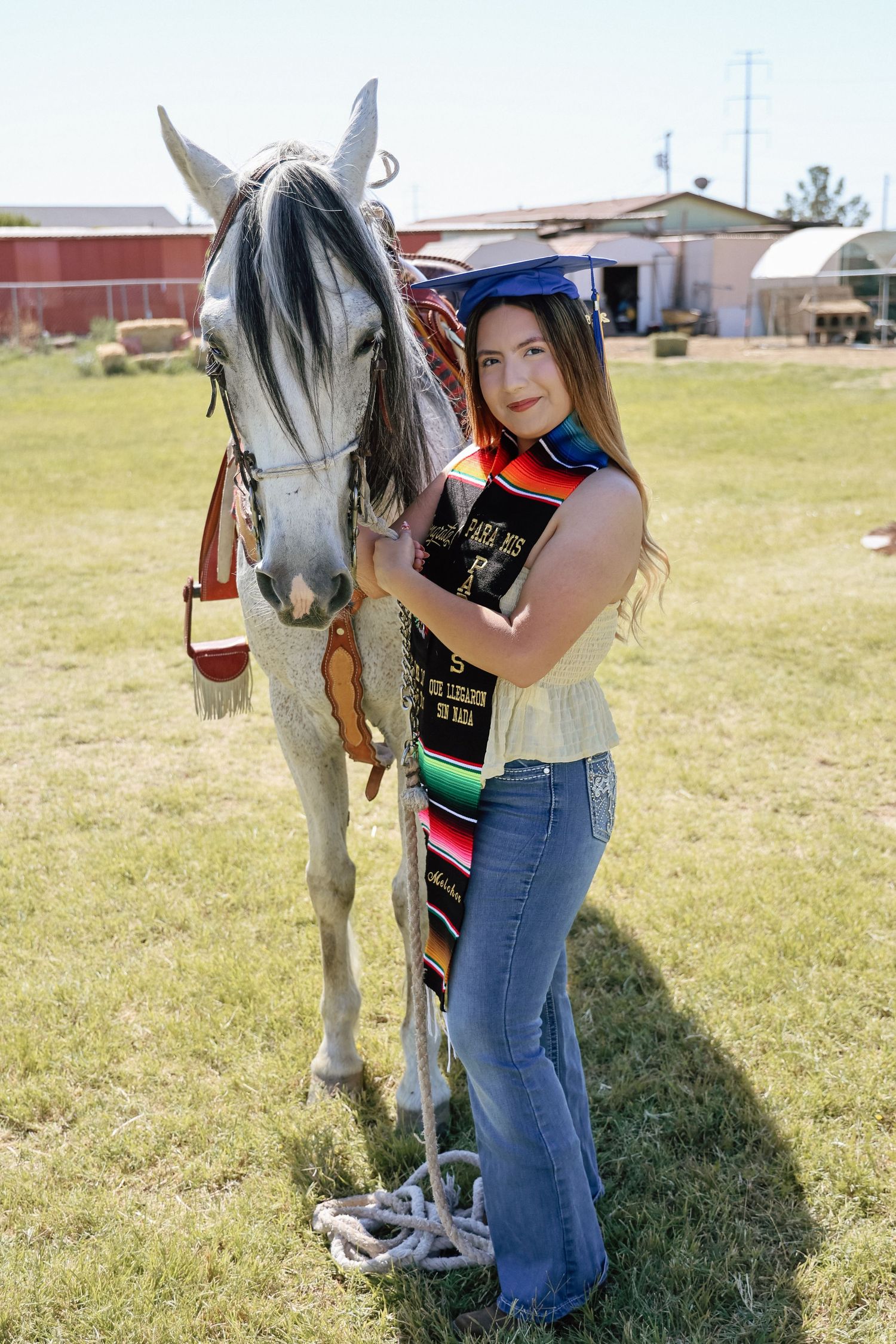 Graduate in cap and colorful sash poses proudly with a white horse on a sunny green lawn.