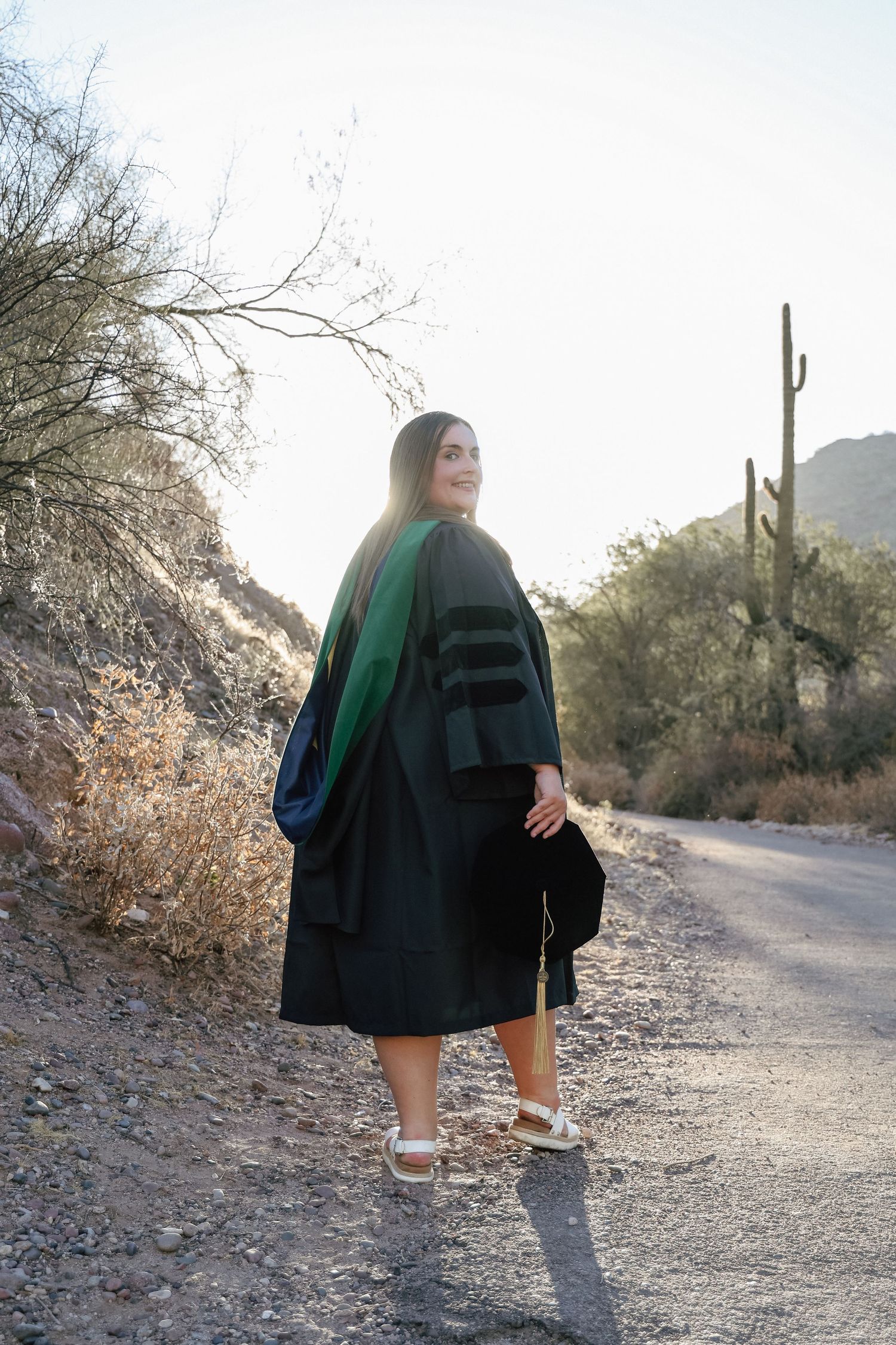 Graduate in black doctoral regalia poses on desert path with saguaro cacti in Arizona landscape.
