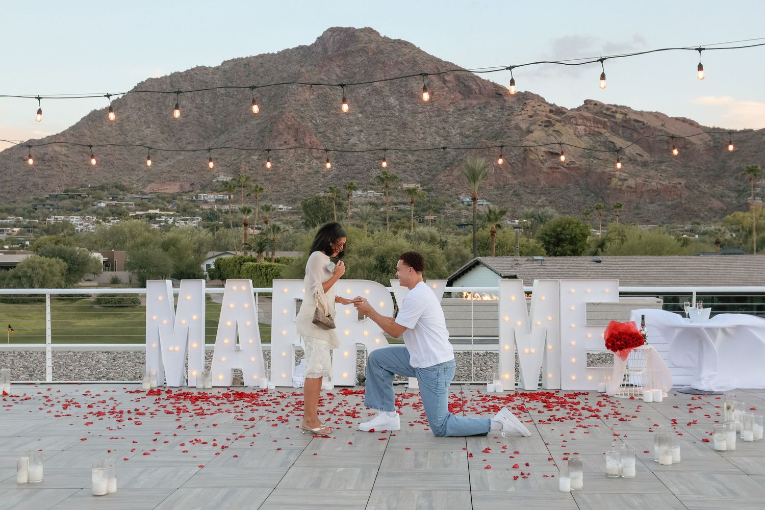 Man proposes on rooftop with MARRY ME letters, rose petals, candles, and mountain backdrop at sunset.