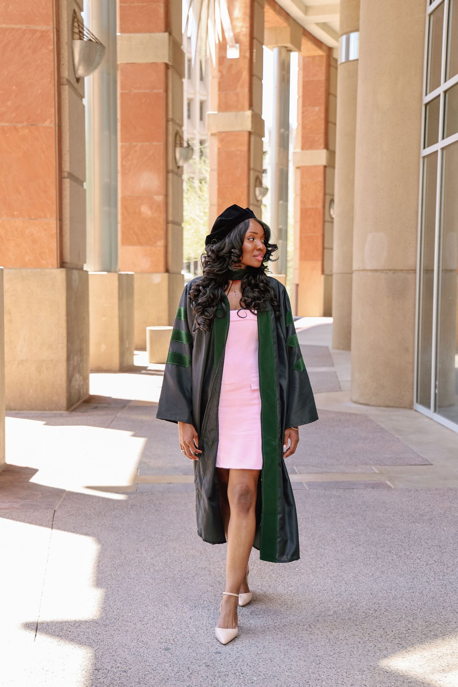 A young woman in a green graduation gown and cap walks confidently in a pink dress and white heels outside a building.