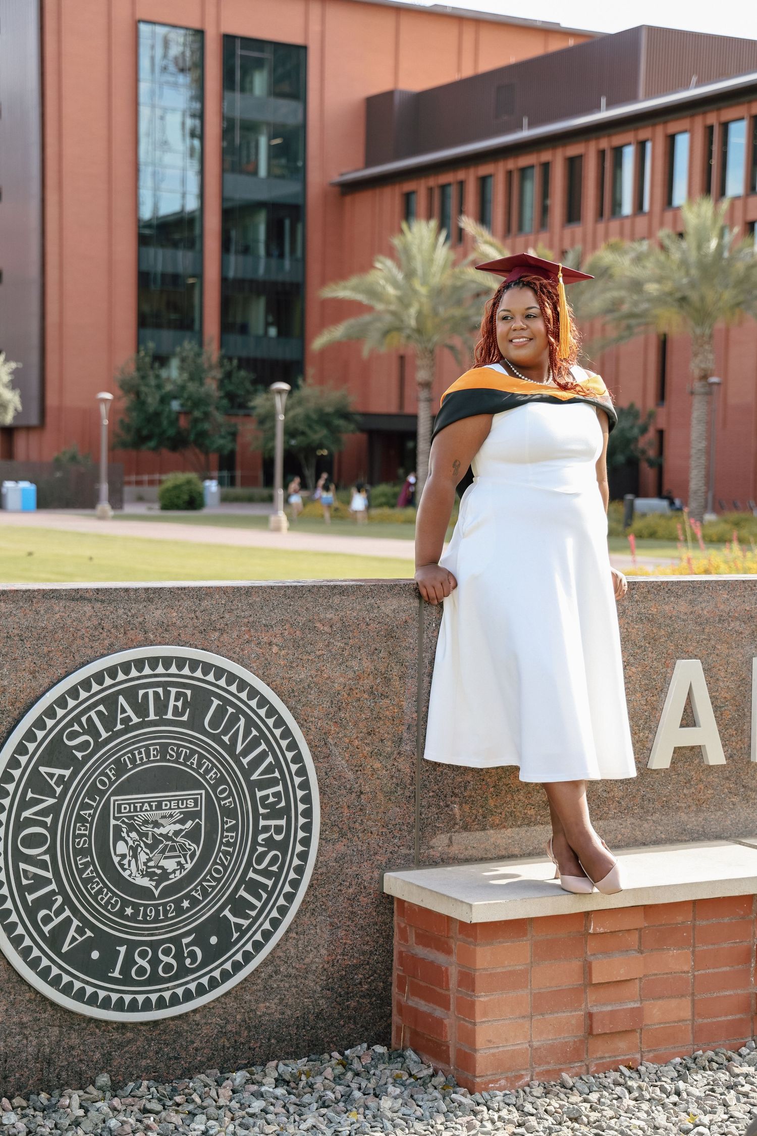 Arizona State University graduate poses proudly in white dress and graduation cap by ASU seal on campus.