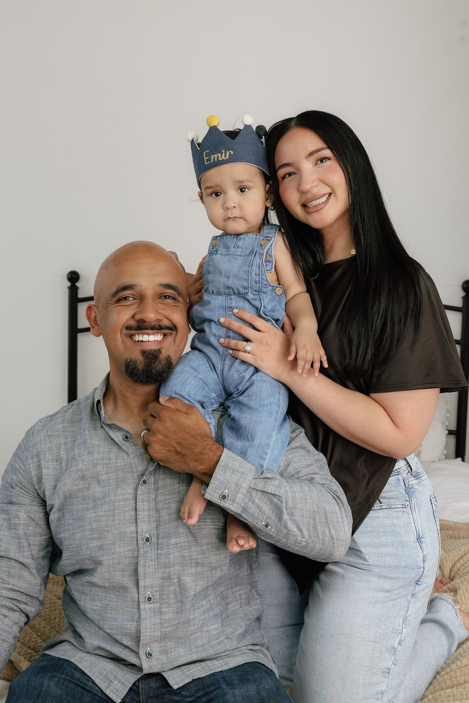 A smiling family with a bald father, long-haired mother, and baby wearing a blue crown and denim overalls posing together.