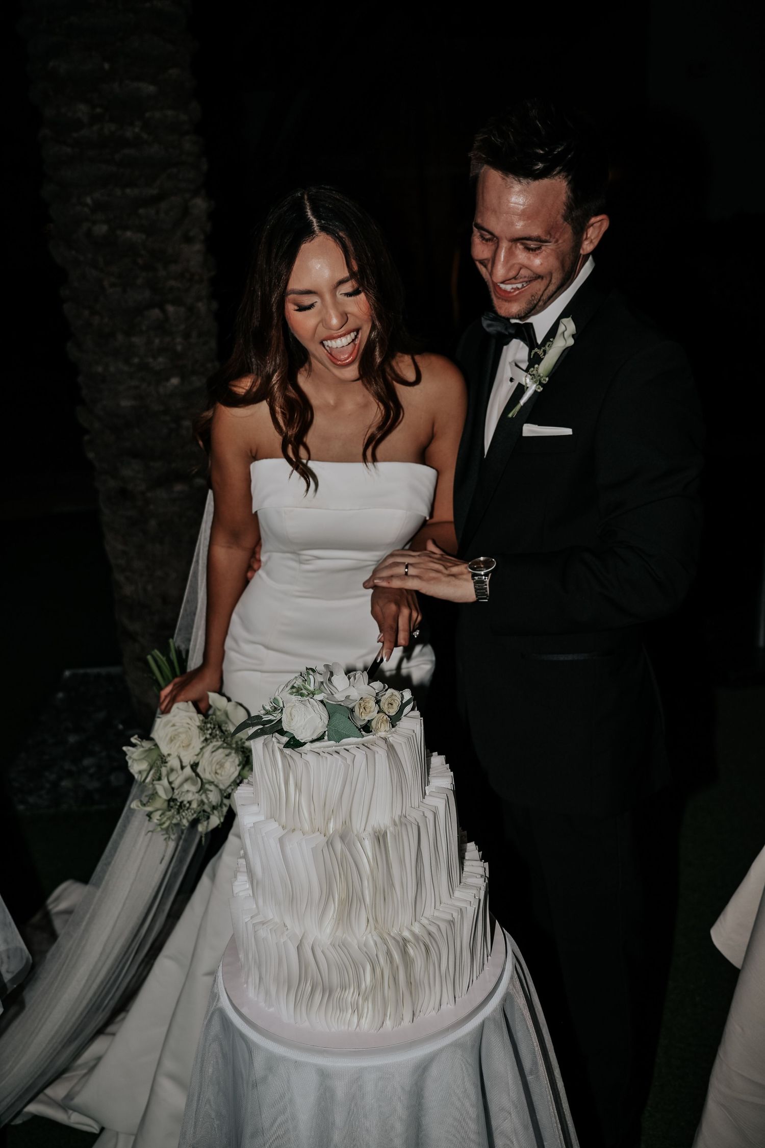Couple cuts into elegant white wedding cake with ruffled tiers in evening setting.
