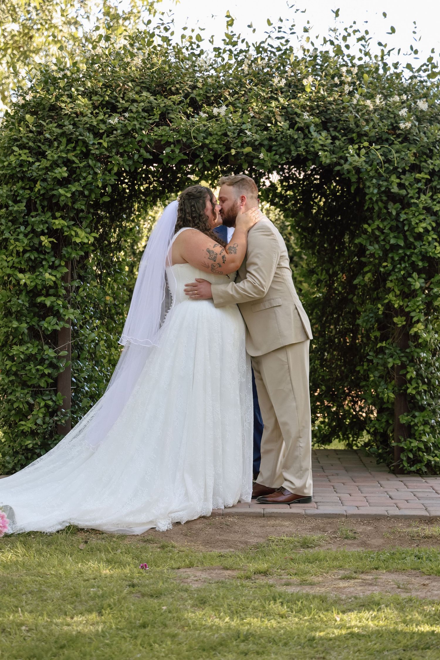 Bride and groom share first kiss under lush green garden arch, bride in white gown with veil, groom in tan suit.