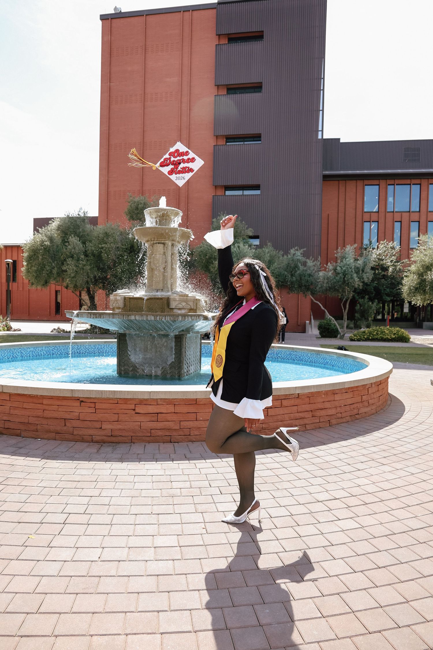 Graduate in cap and gown tossing mortarboard by a fountain outside a brick university building.