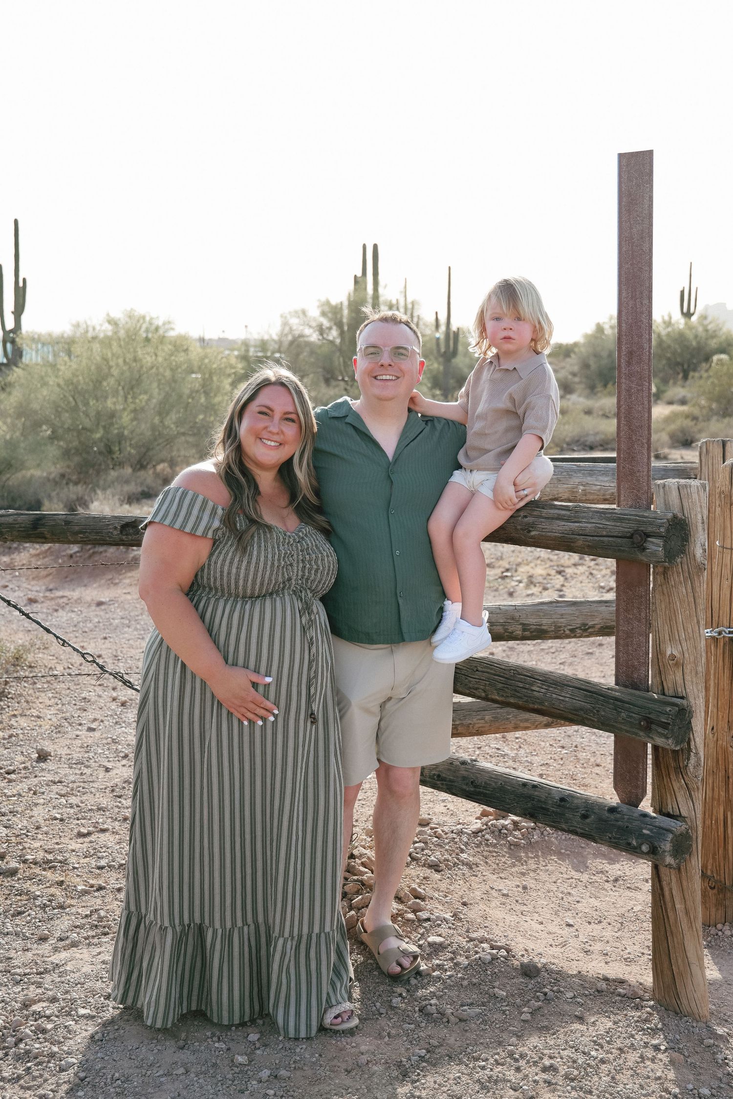 Pregnant woman in green striped dress poses with man and toddler by rustic fence in desert landscape.