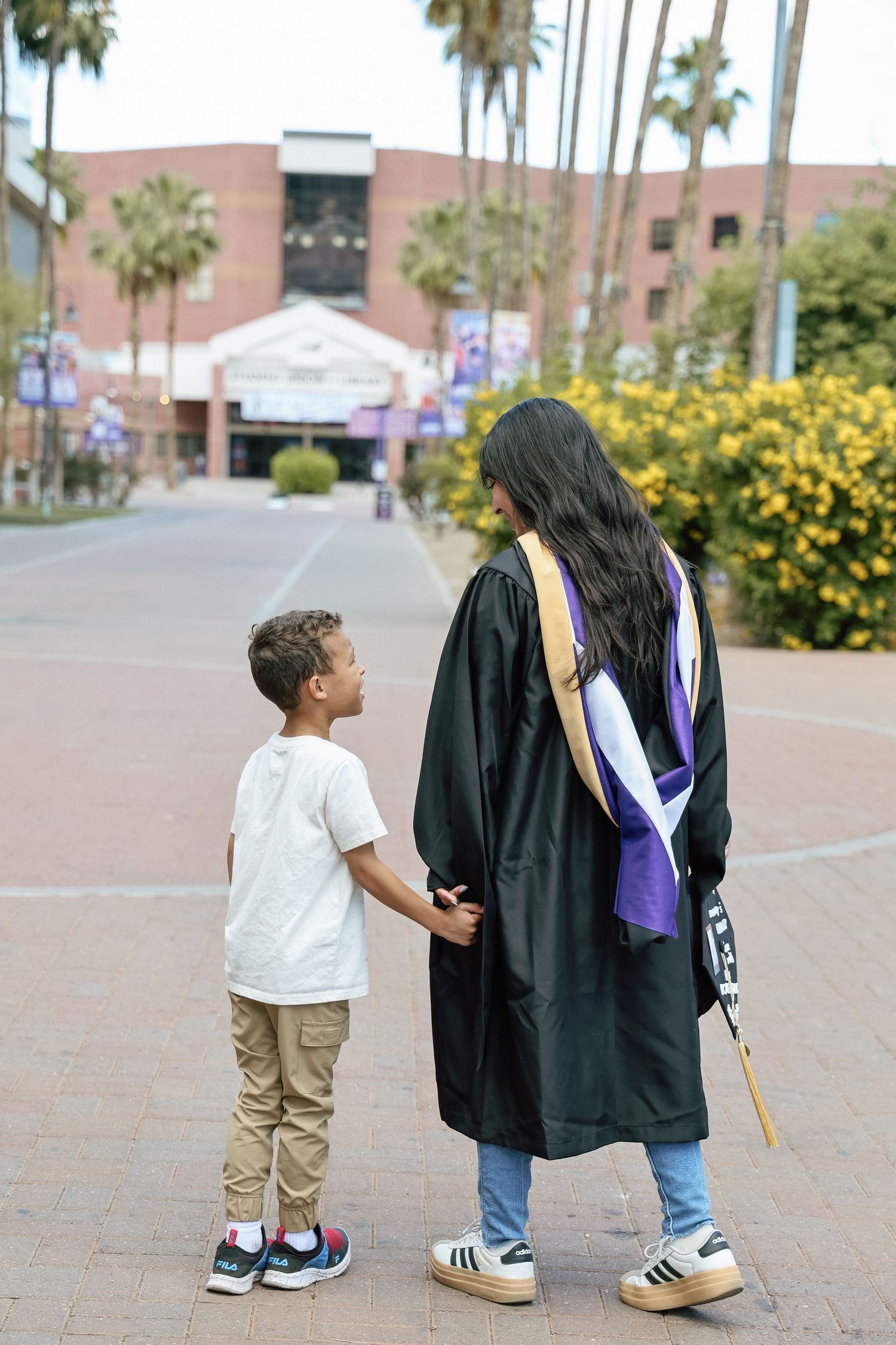A graduate in black cap and gown holds hands with a young boy on a university campus lined with palm trees.
