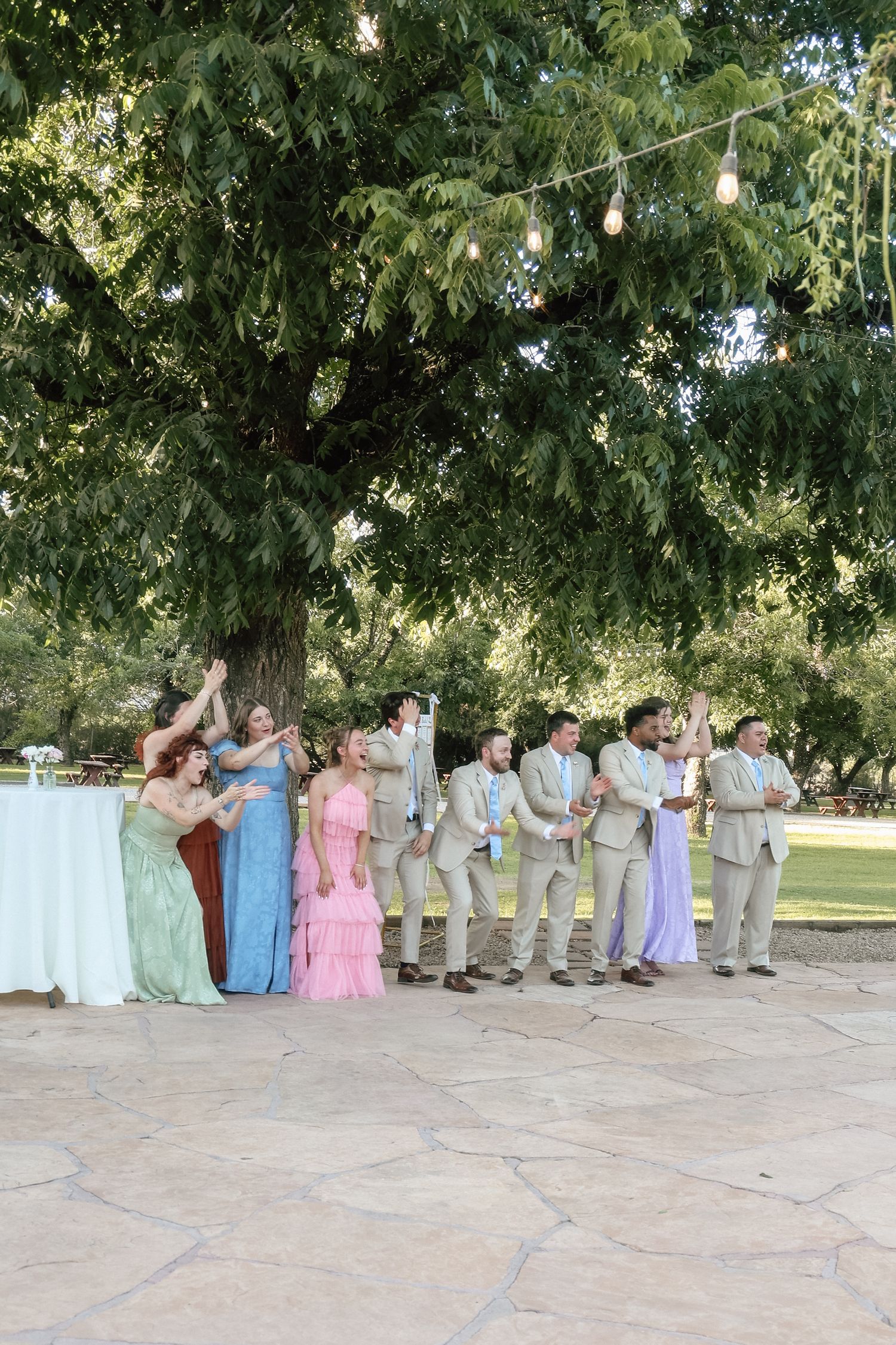Wedding party plays lawn games under a large tree with string lights at an outdoor reception.