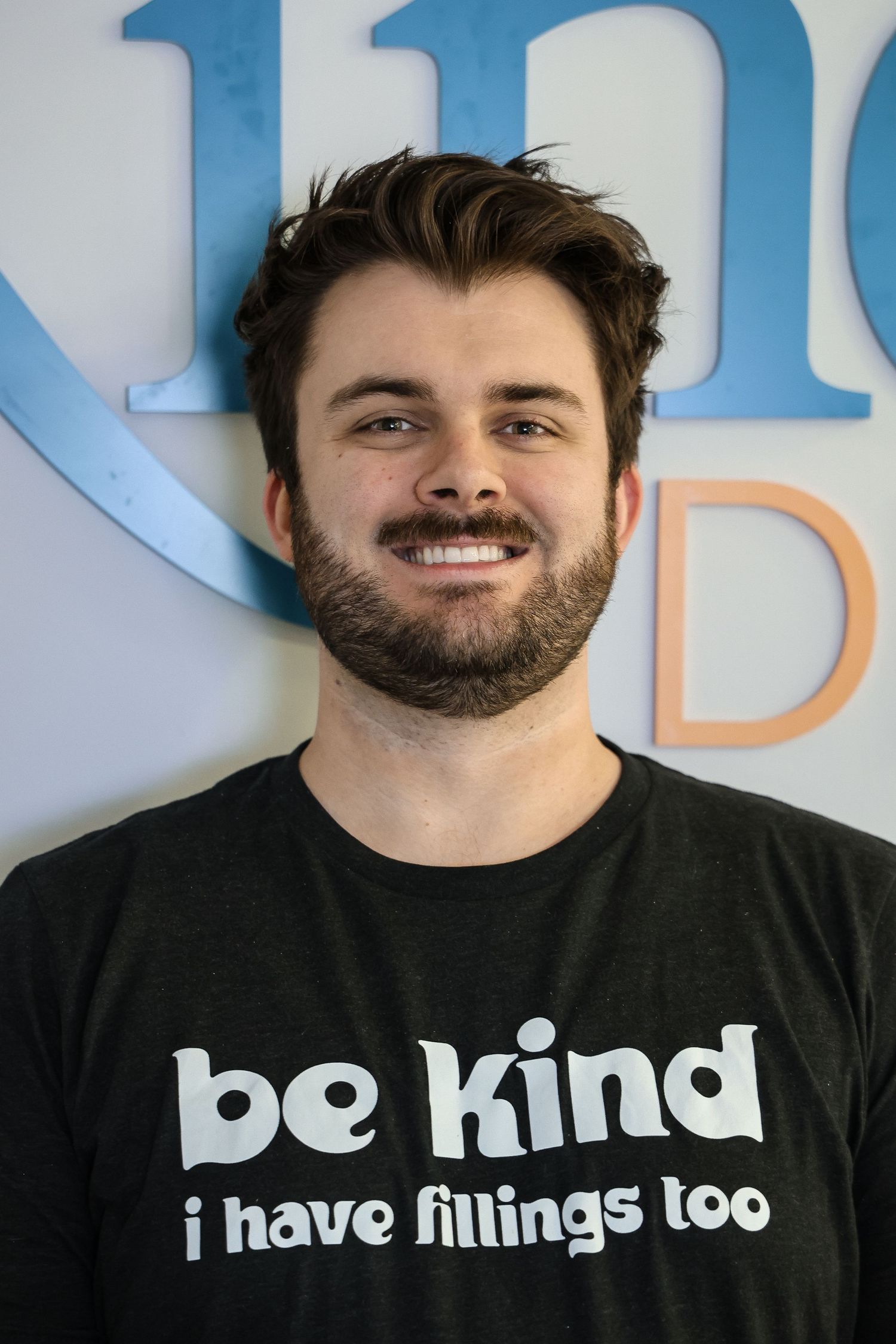 Smiling young man with beard wearing a black 'be kind i have fillings too' t-shirt standing indoors.