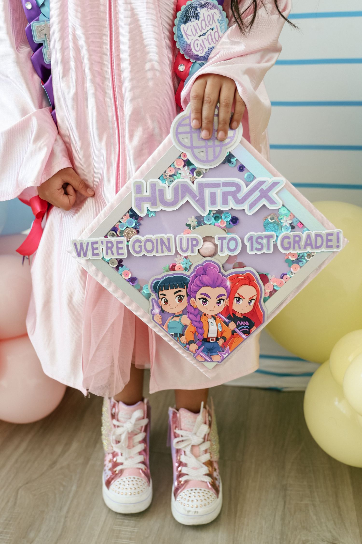 Child in pink graduation gown holding a decorative cap reading Huntryx We're Going Up to 1st Grade.