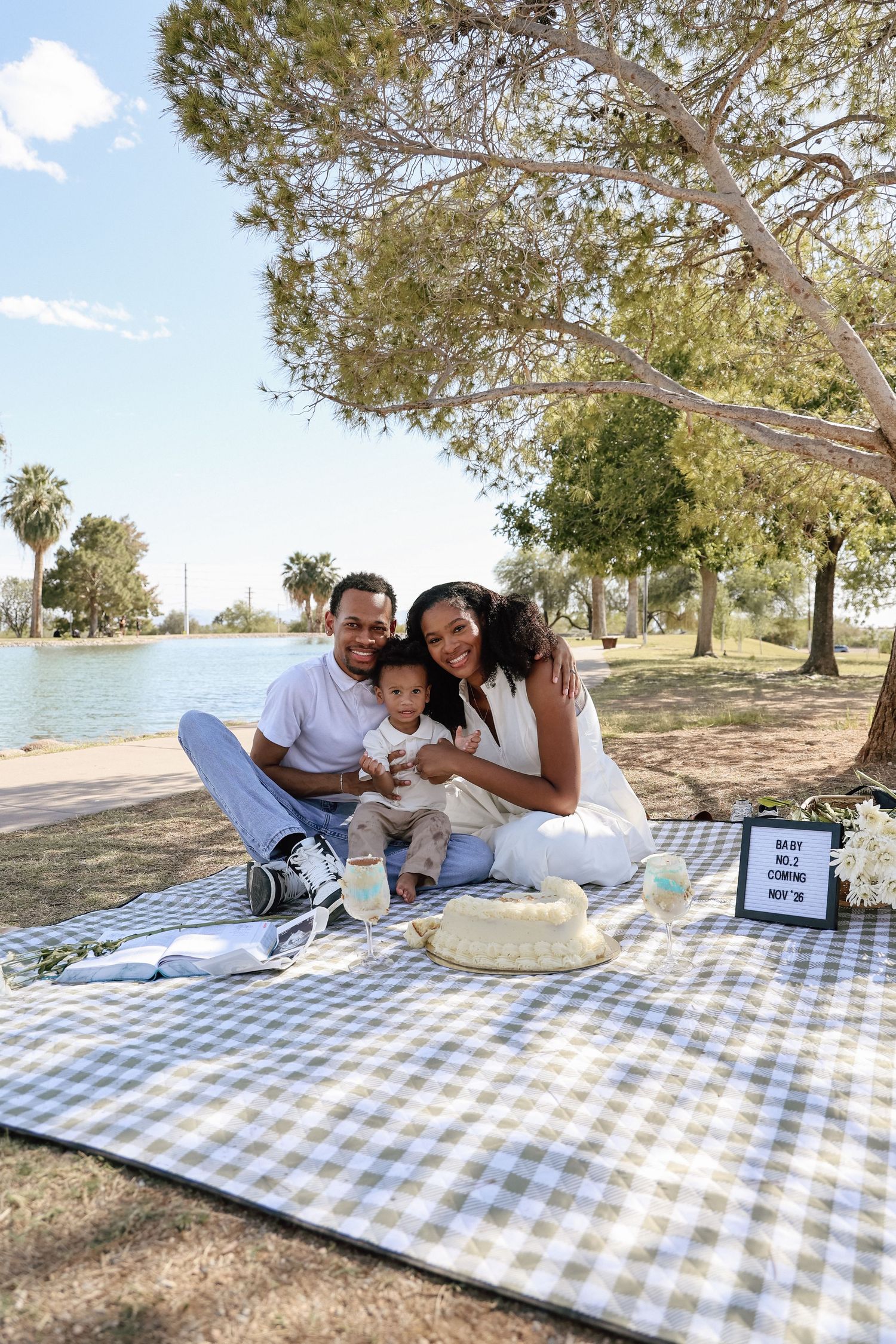 Family of three celebrating baby's first birthday with a cake at a park picnic by the lake.