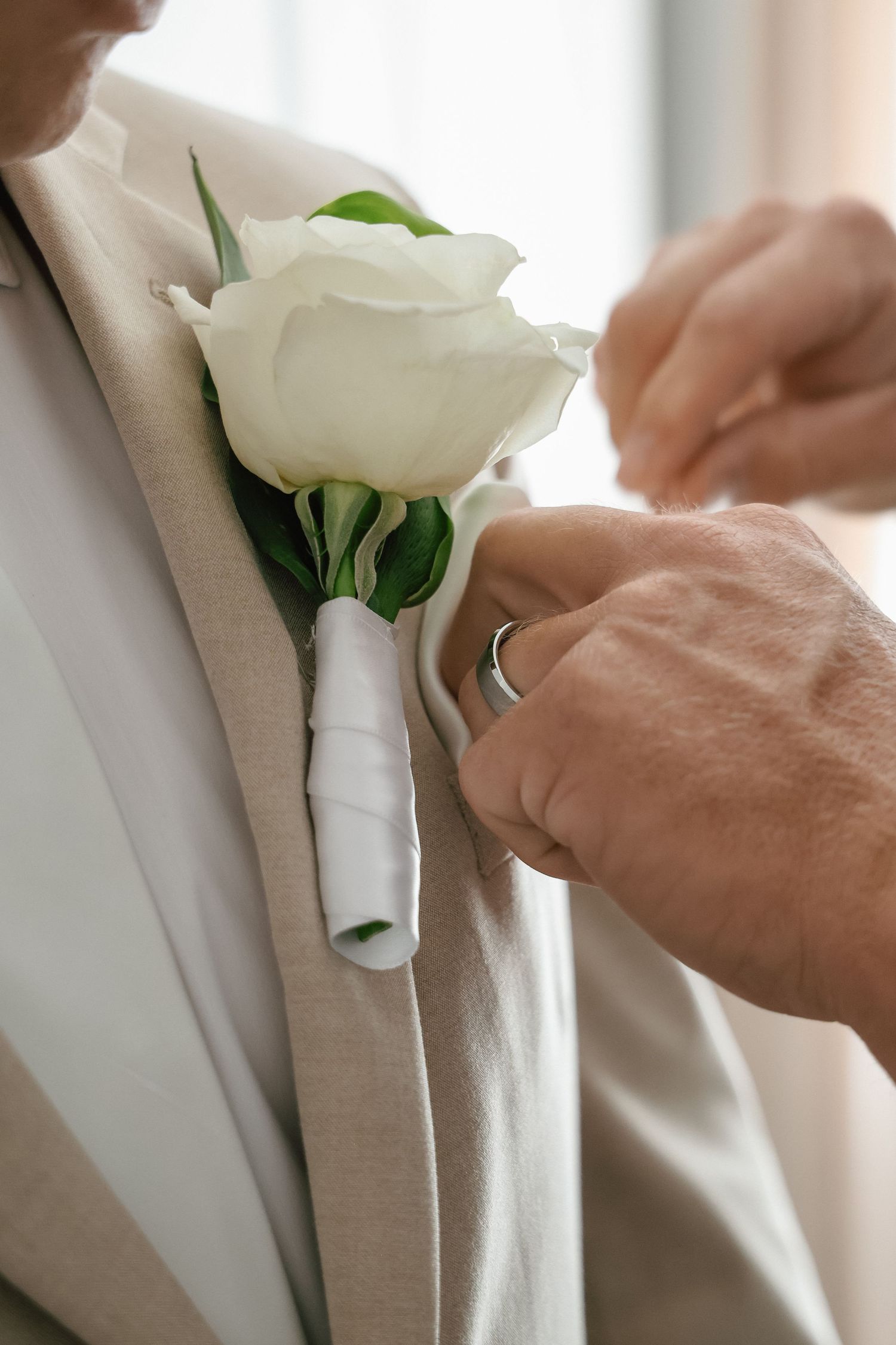 Hands pinning a white rose boutonniere onto a beige suit lapel for a wedding ceremony.