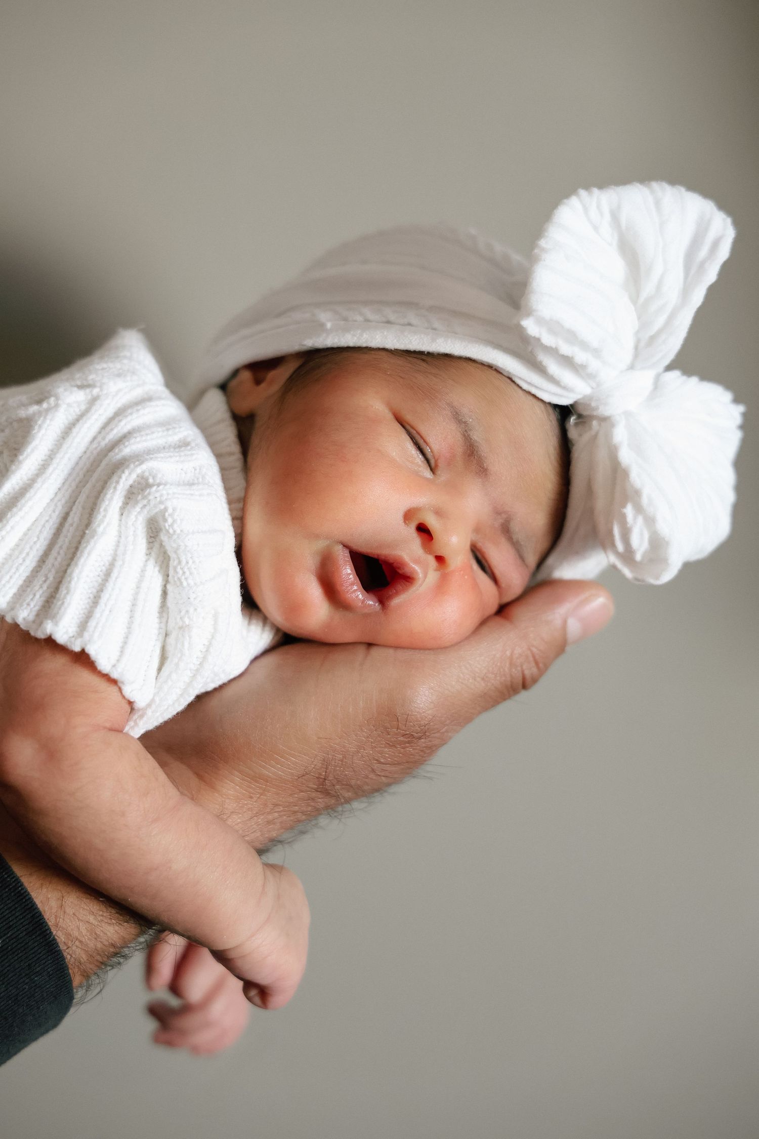 Newborn baby sleeping peacefully in a parent's hand, wearing a white knit outfit and bow hat.