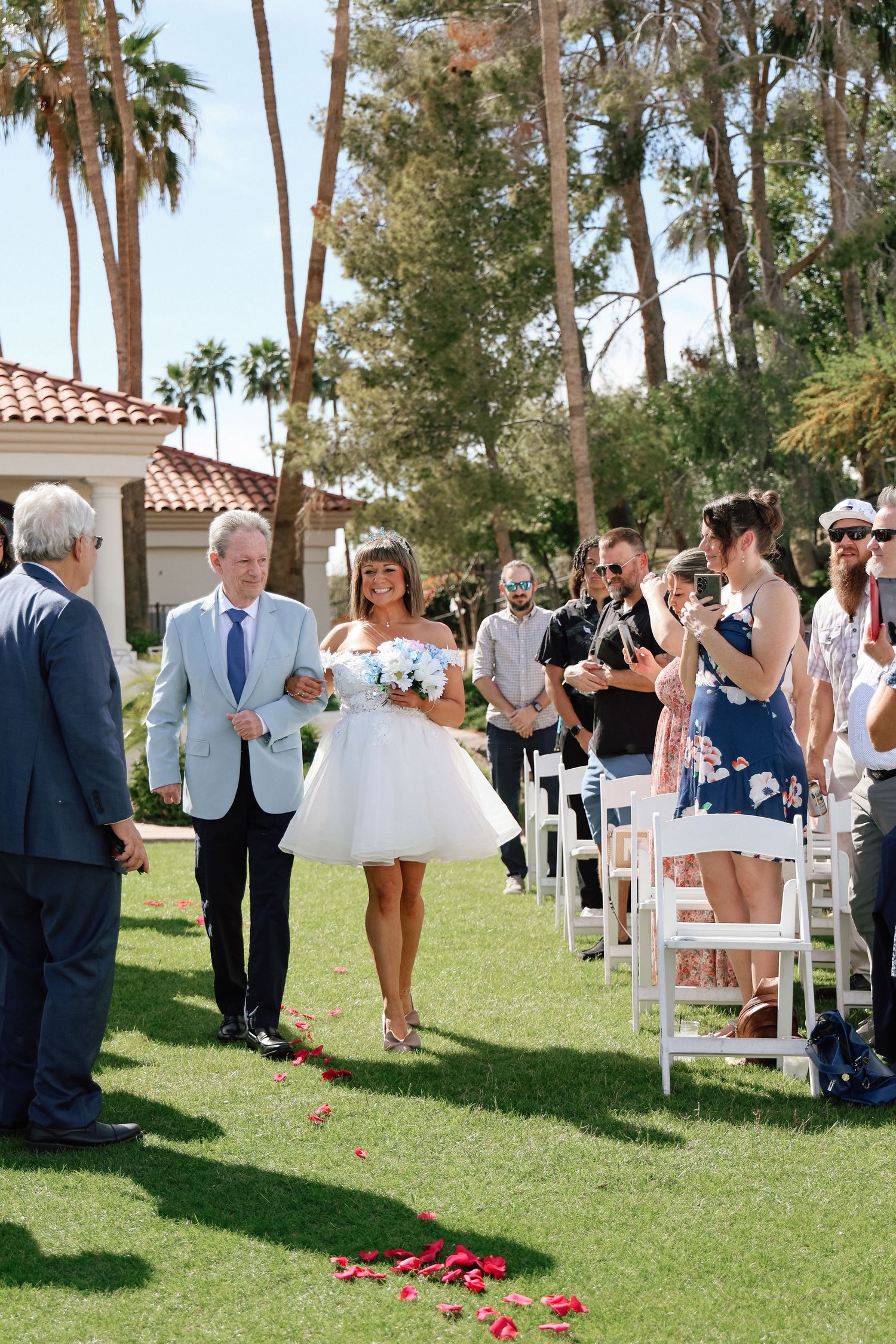 Bride in short white dress walks down outdoor aisle with escort, guests cheering, palm trees and rose petals on green lawn.