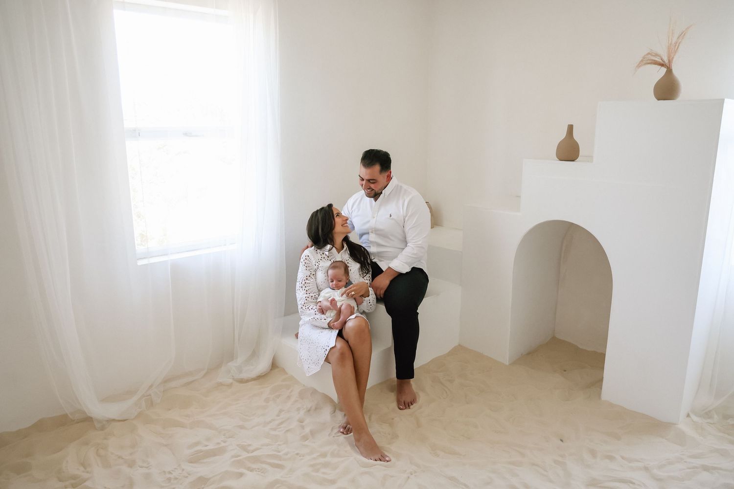 A couple sits together in a bright white studio, tenderly holding their newborn baby in a serene family portrait.