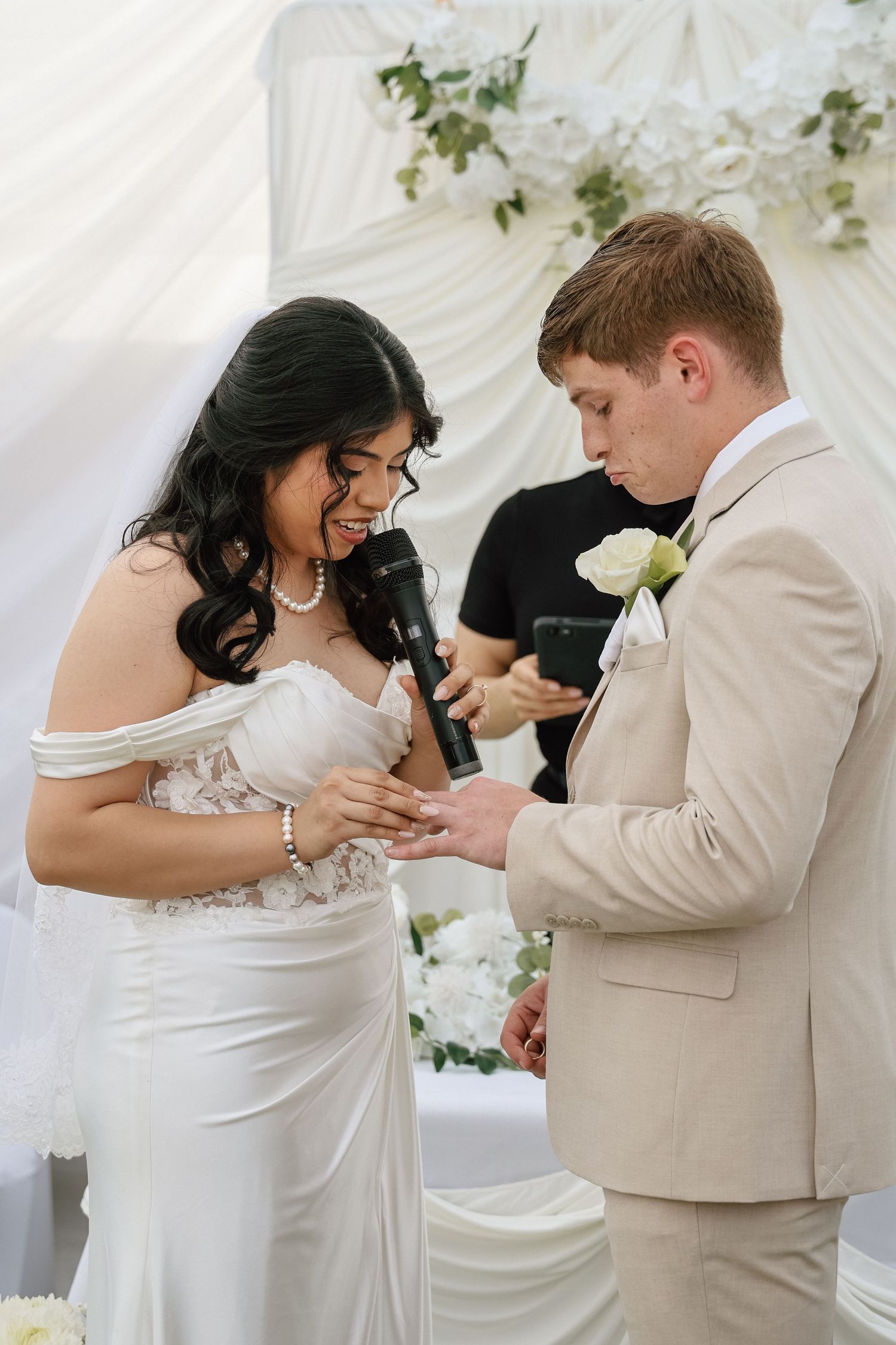 Bride and groom exchanging rings at elegant white wedding ceremony, groom in beige suit, bride in off-shoulder lace gown.