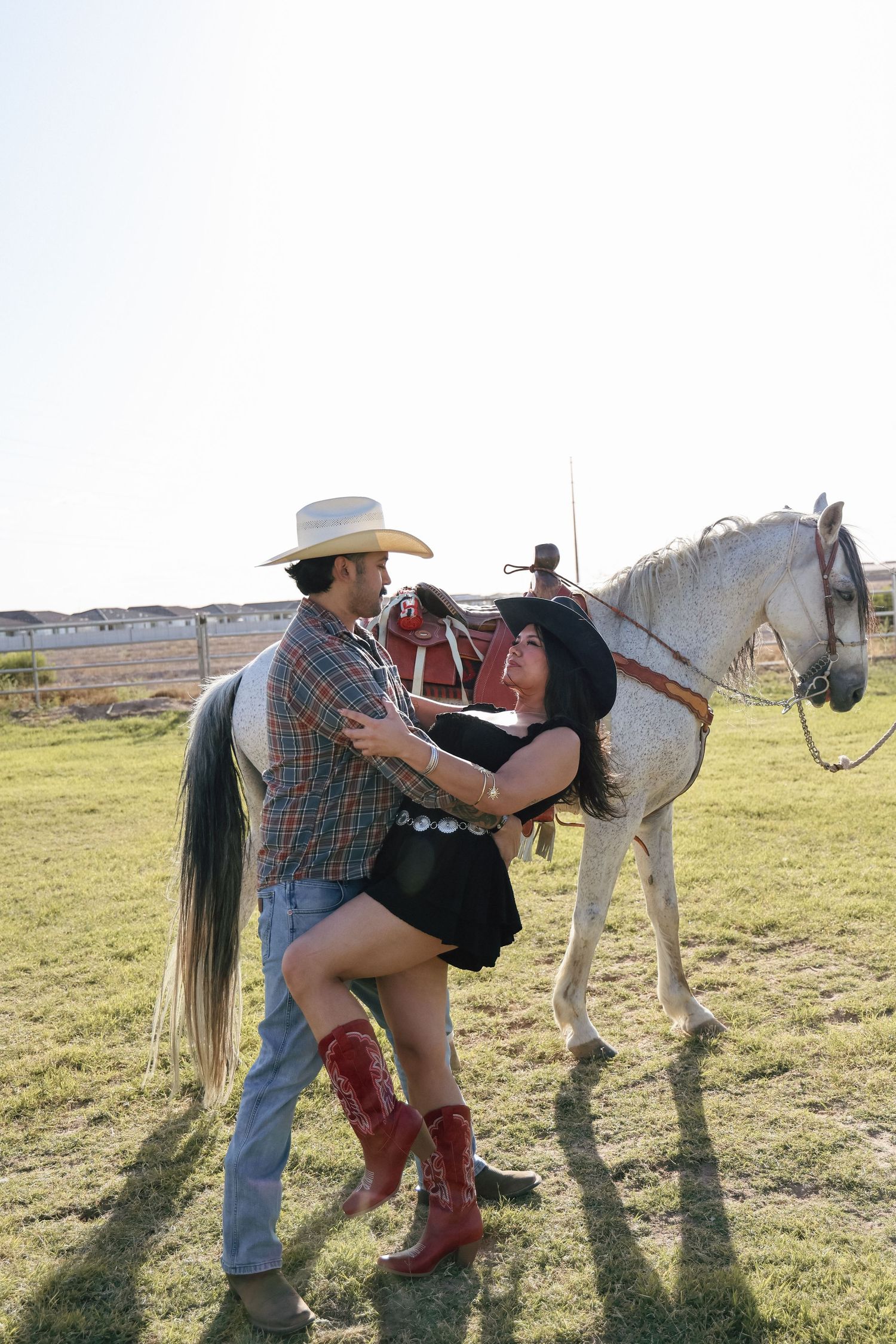 Couple in cowboy hats embracing near a white horse on a sunny ranch, woman wearing red boots and black dress.