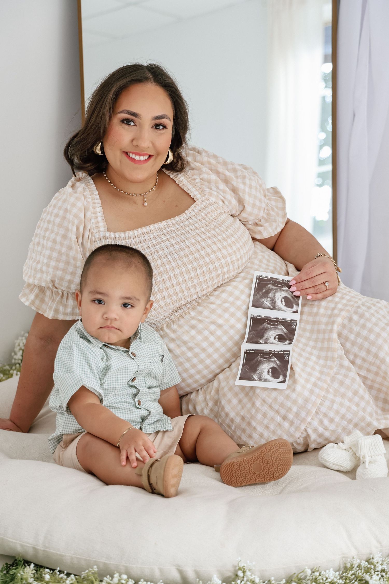 Pregnant woman in gingham dress holds ultrasound photos beside her baby boy on a white cushion.