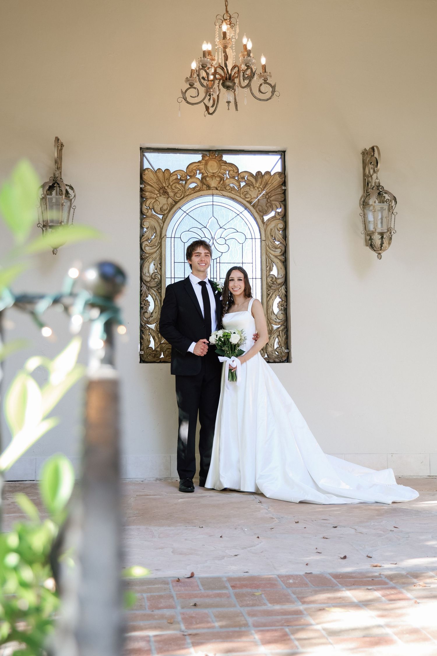 Bride and groom posing elegantly in front of ornate stained glass window with chandelier overhead on wedding day.