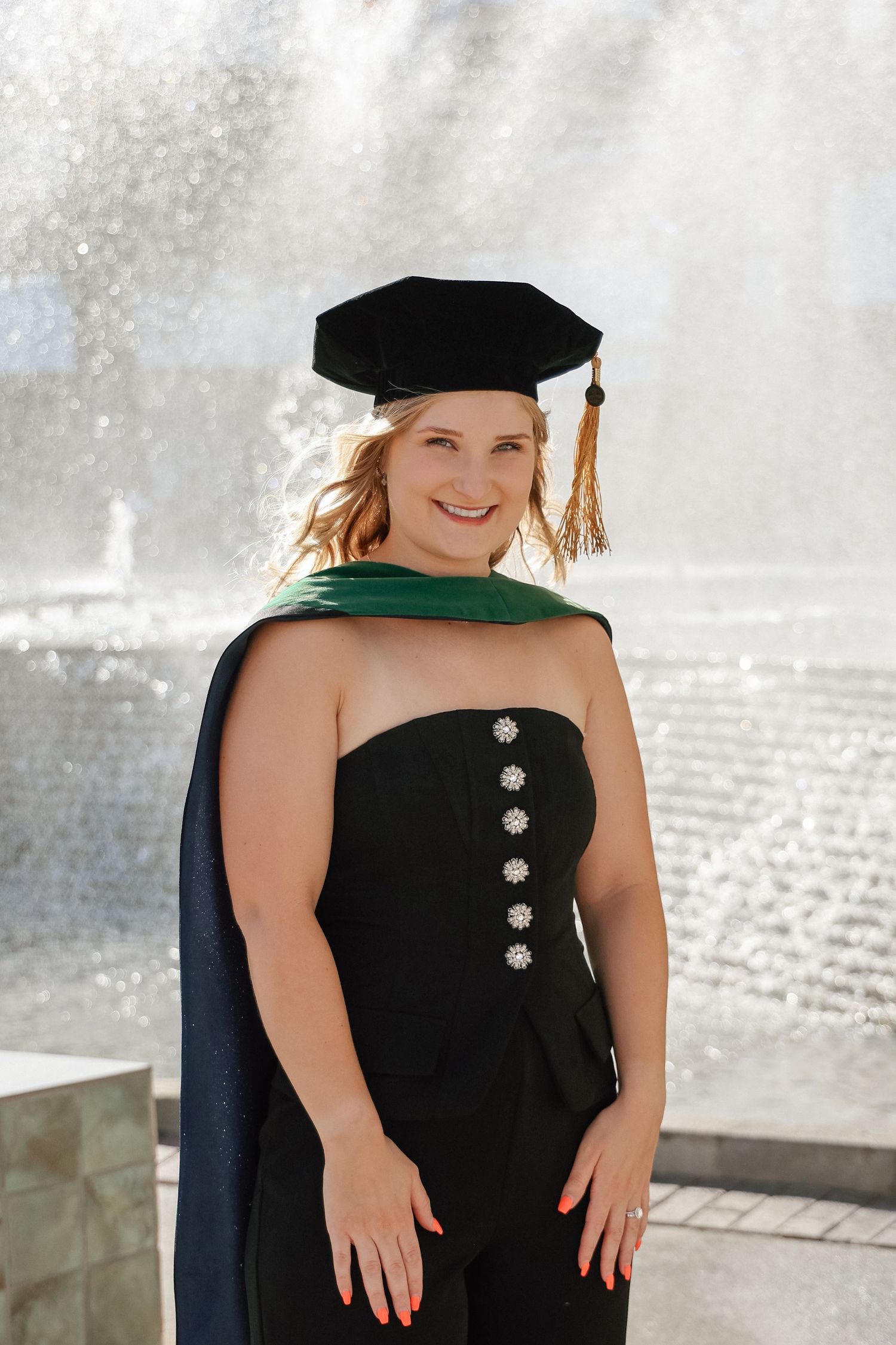 Smiling graduate woman in black cap and gown with green hood posing in front of a fountain.