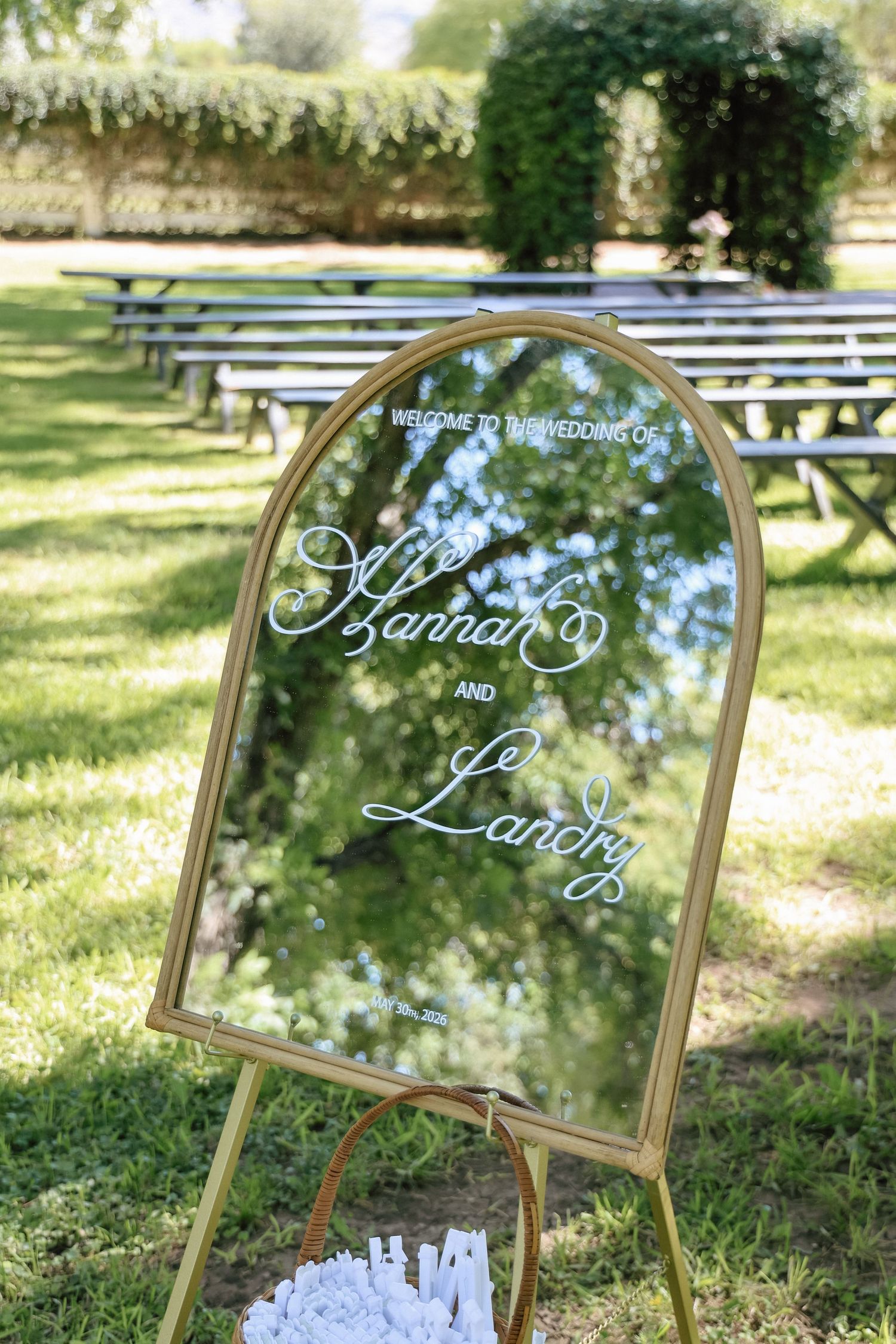 Gold-framed arch mirror wedding welcome sign for Hannah and Landry displayed on easel outdoors among green trees.