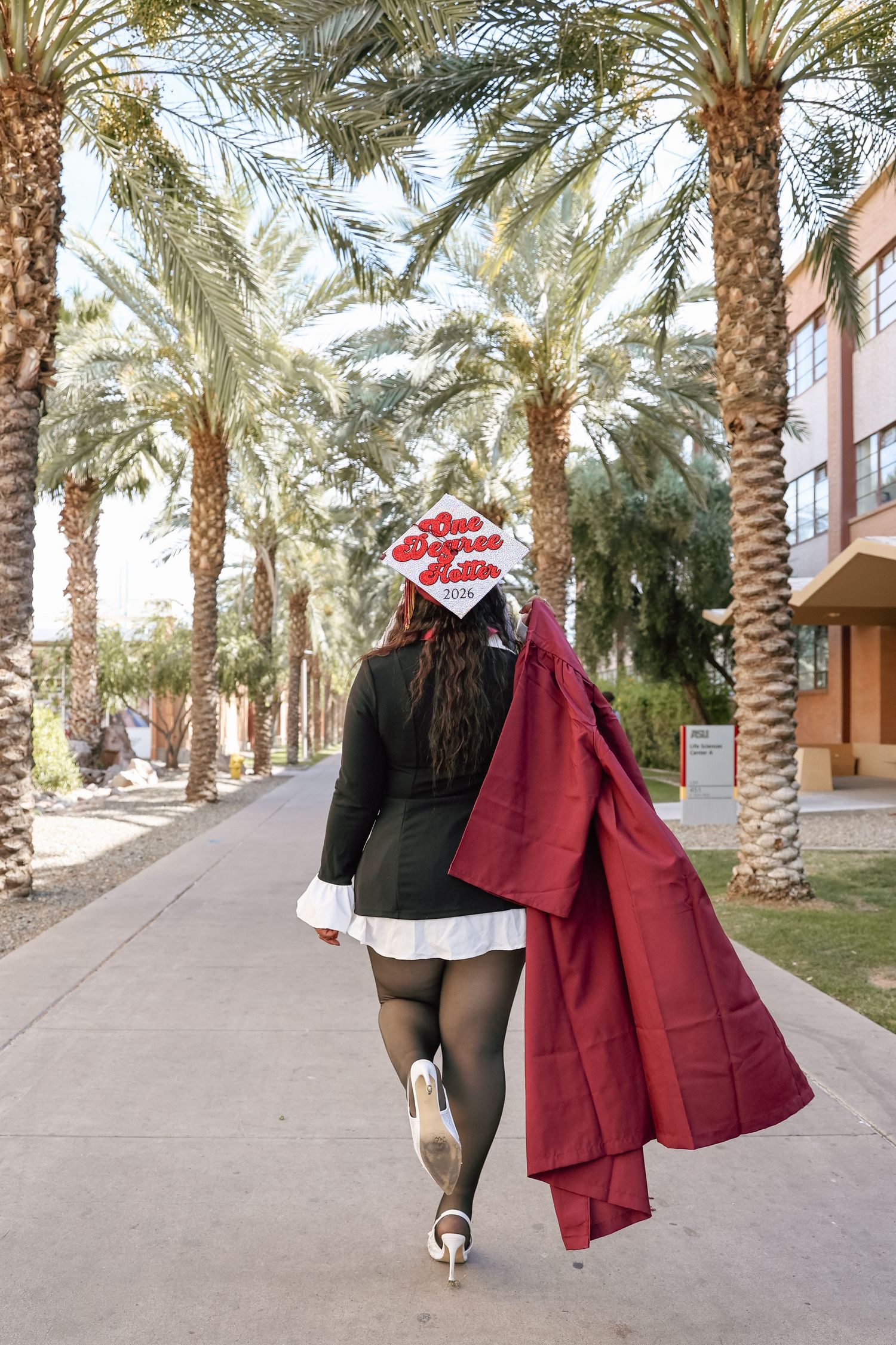 Graduate walks away in cap and gown, holding crimson robe, flanked by palm trees on sunny campus path.