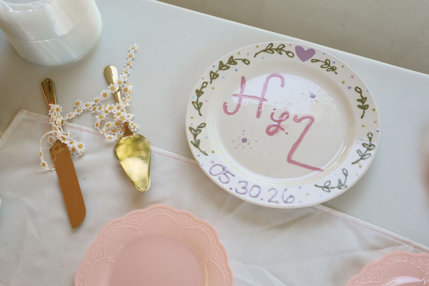 Hand-painted wedding plate with initials and date 05.30.26, displayed beside gold cake servers and pink plates.