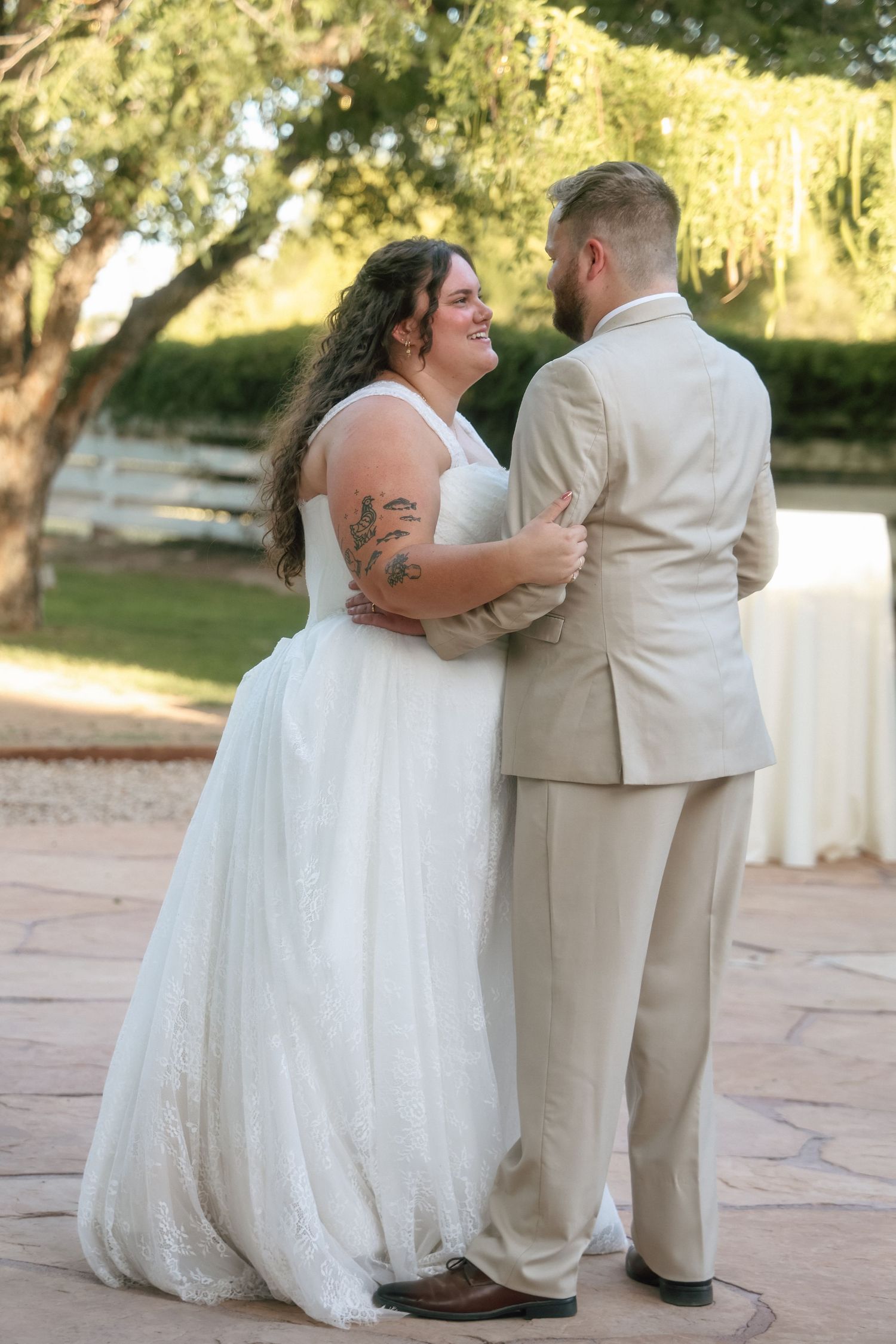 Bride in white lace gown with tattoos shares first dance with groom in tan suit at outdoor wedding reception.