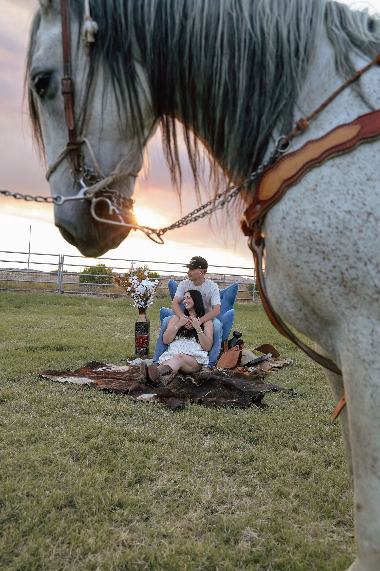 Couple sitting on blanket at sunset with a gray dapple horse in foreground on a ranch.