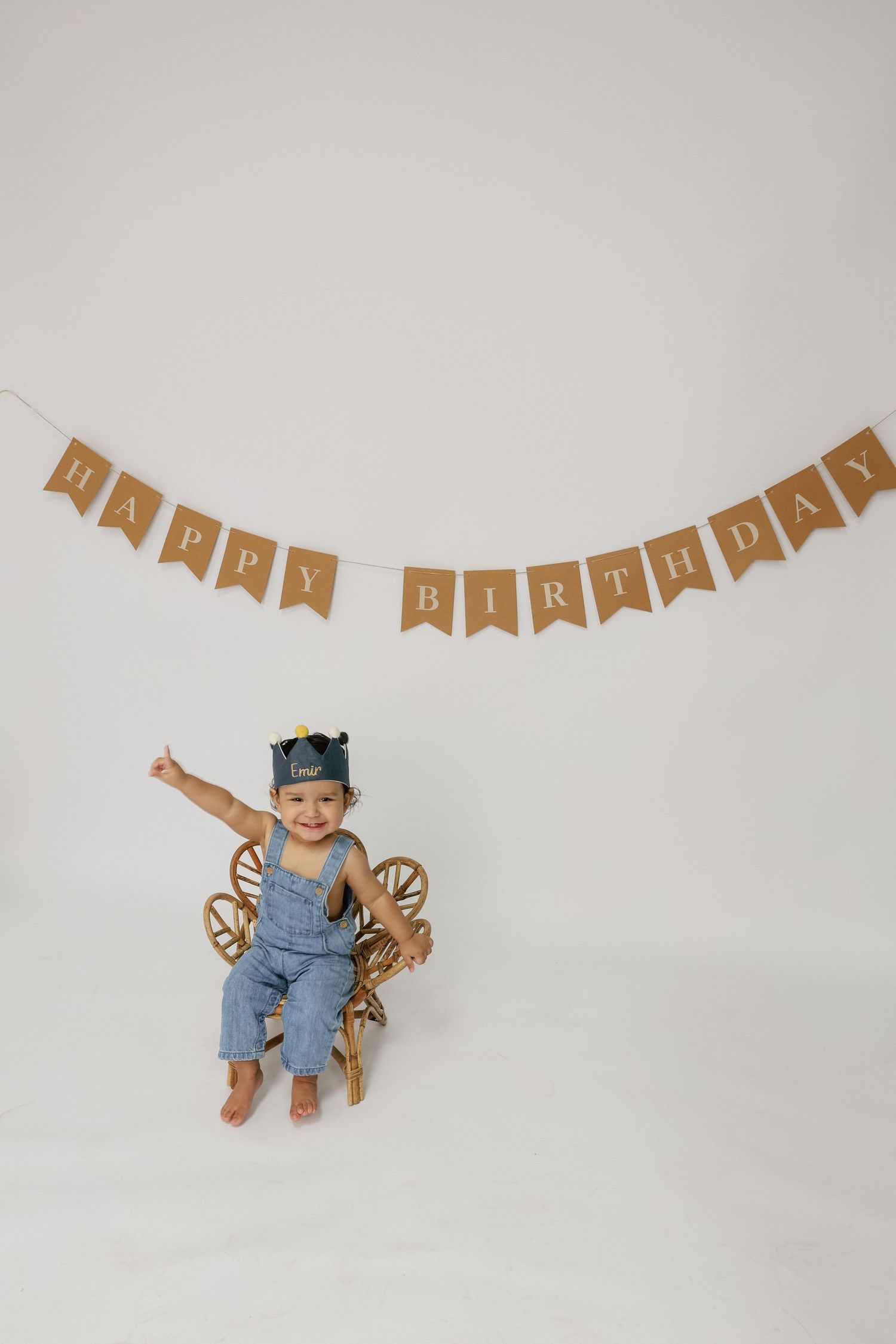 Smiling toddler in denim overalls and crown hat sits on wooden chair under Happy Birthday bunting banner.