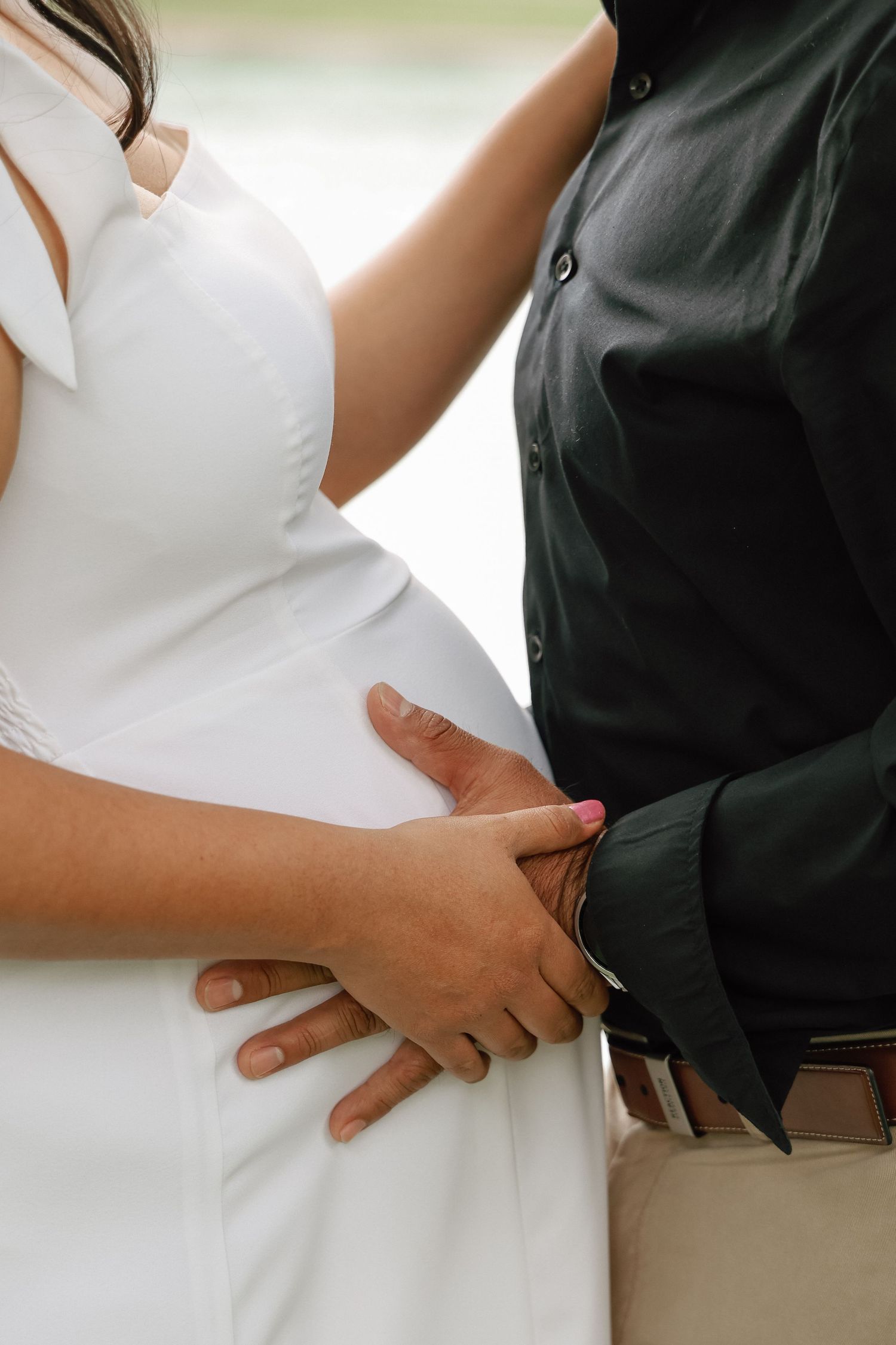 Couple holding hands tenderly, pregnant woman in white dress with partner in black shirt during maternity photoshoot.