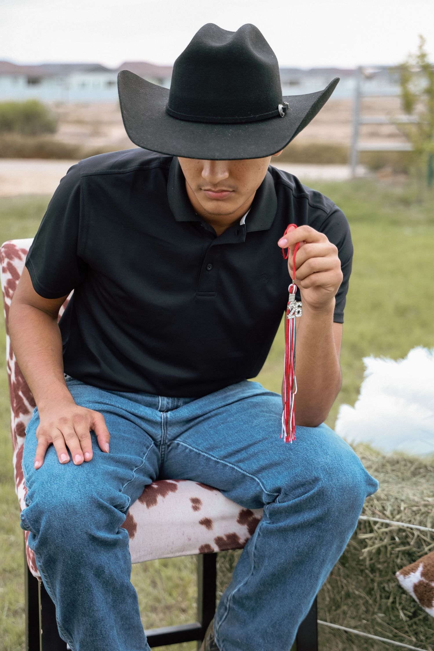 A cowboy in a black hat and jeans sits outdoors holding a red tassel accessory on a cowhide chair.