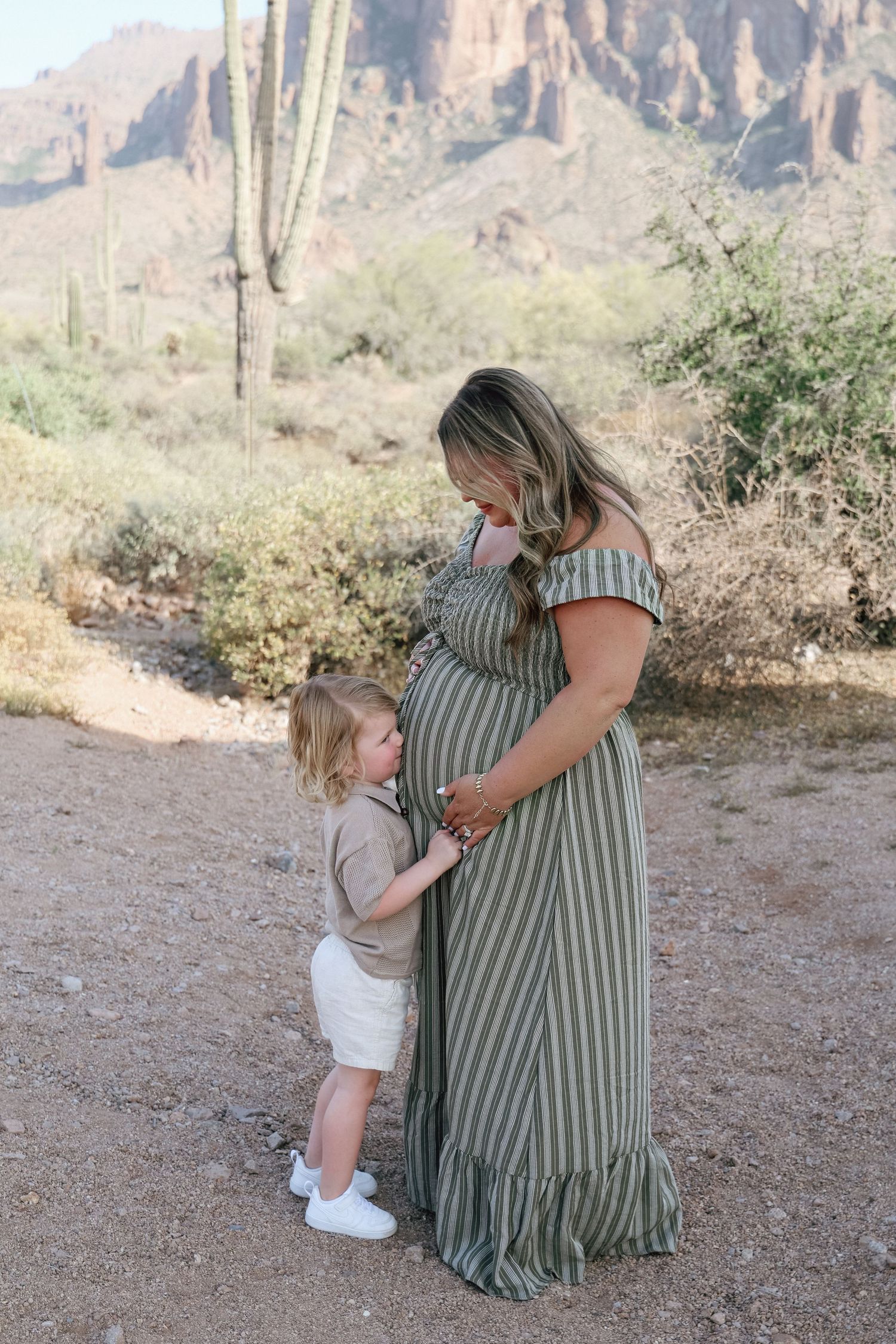 Pregnant woman in striped maxi dress with toddler touching her belly in Arizona desert landscape.