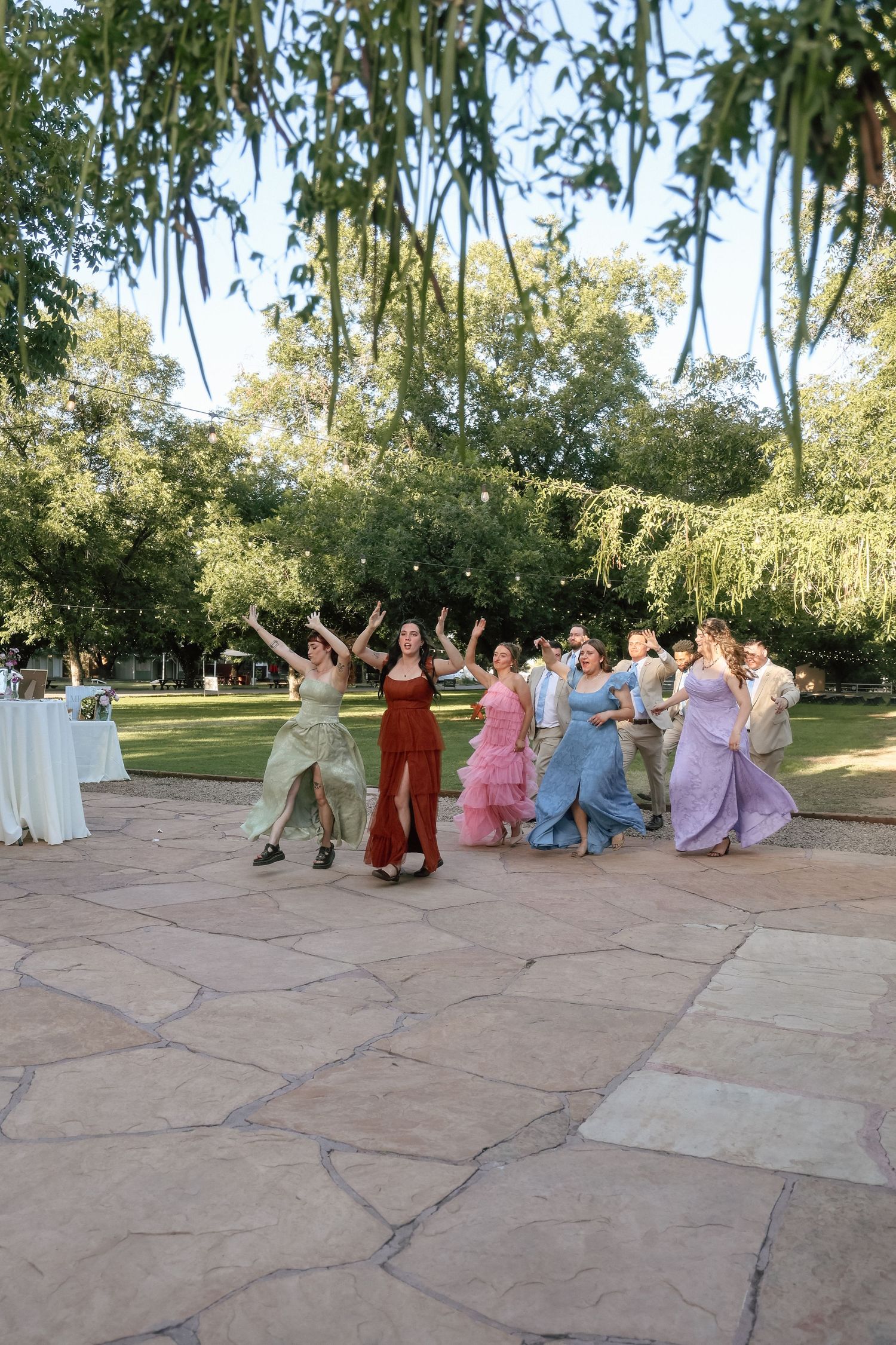 Group of elegantly dressed guests dancing outdoors on stone patio at garden wedding reception.