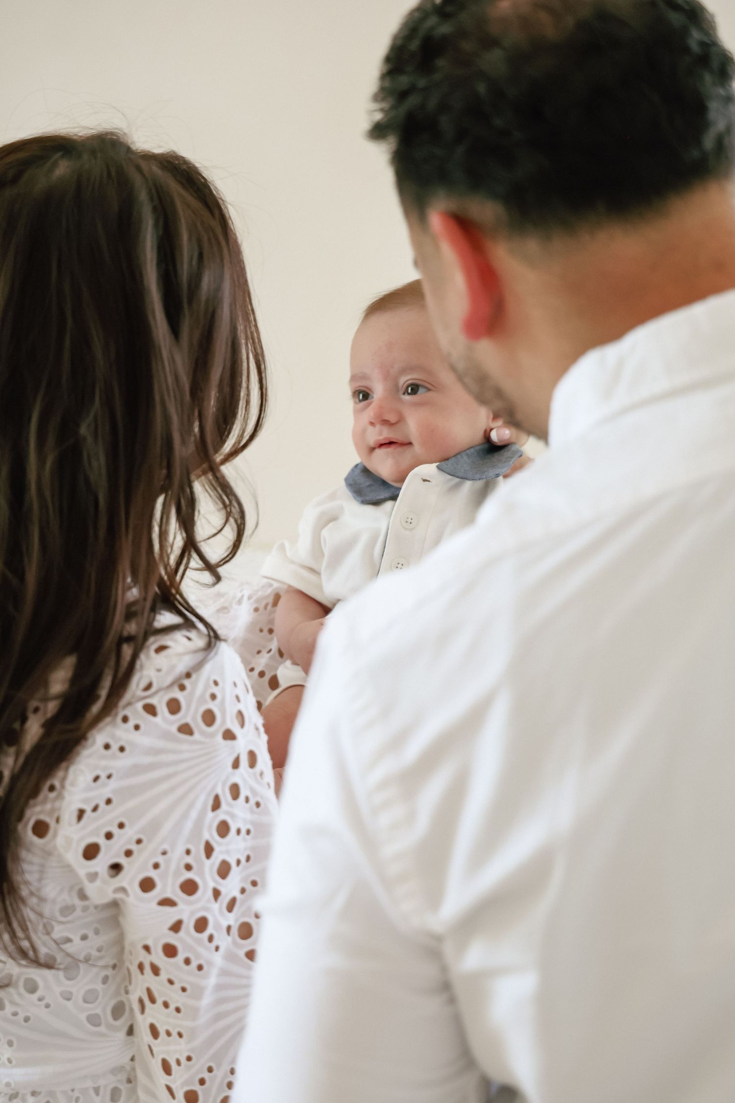 Parents in white clothing lovingly holding their smiling baby dressed in a white outfit with a gray bow tie.