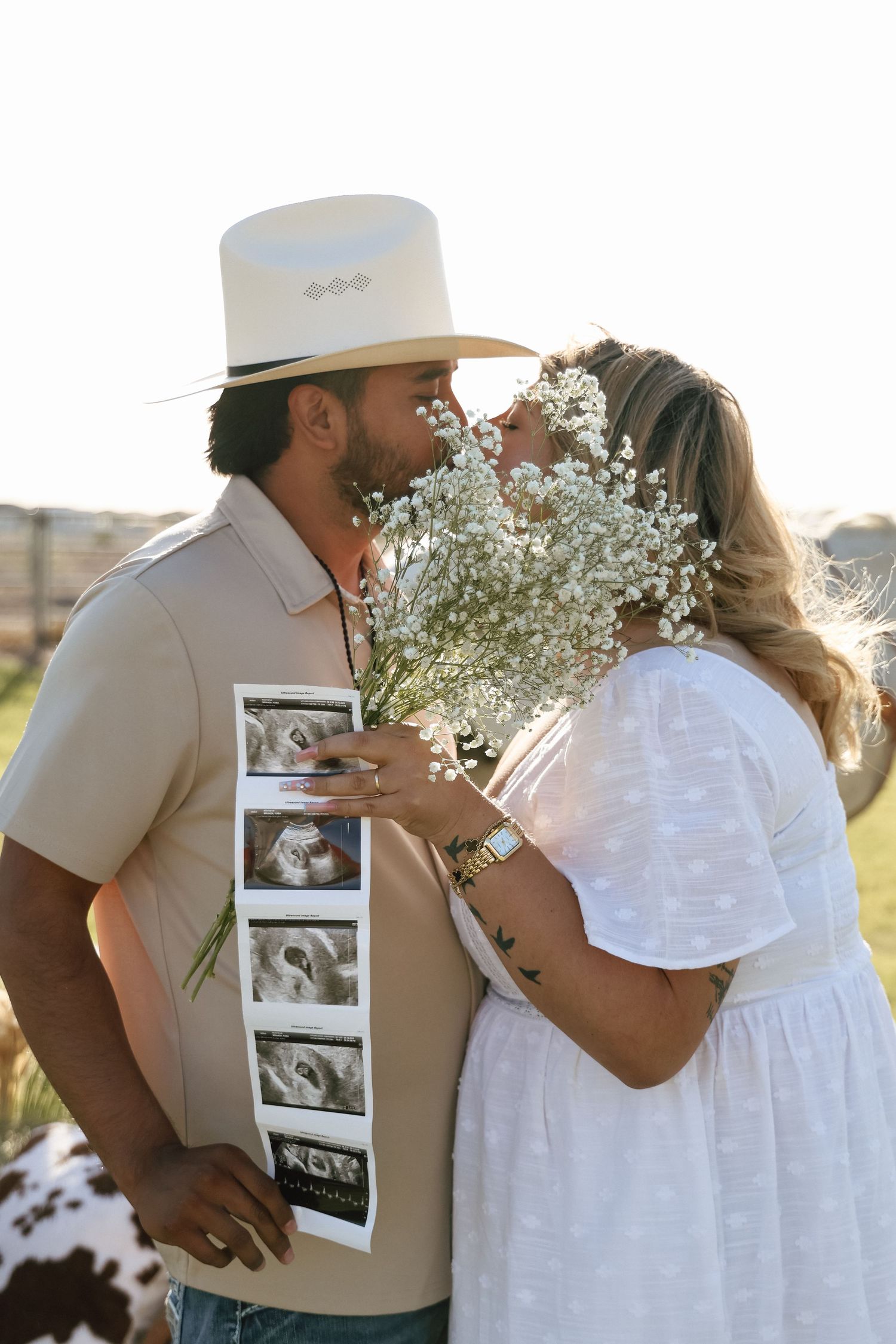 Couple kissing behind baby's breath flowers, man holding ultrasound photos in cowboy hat, pregnancy announcement outdoors.
