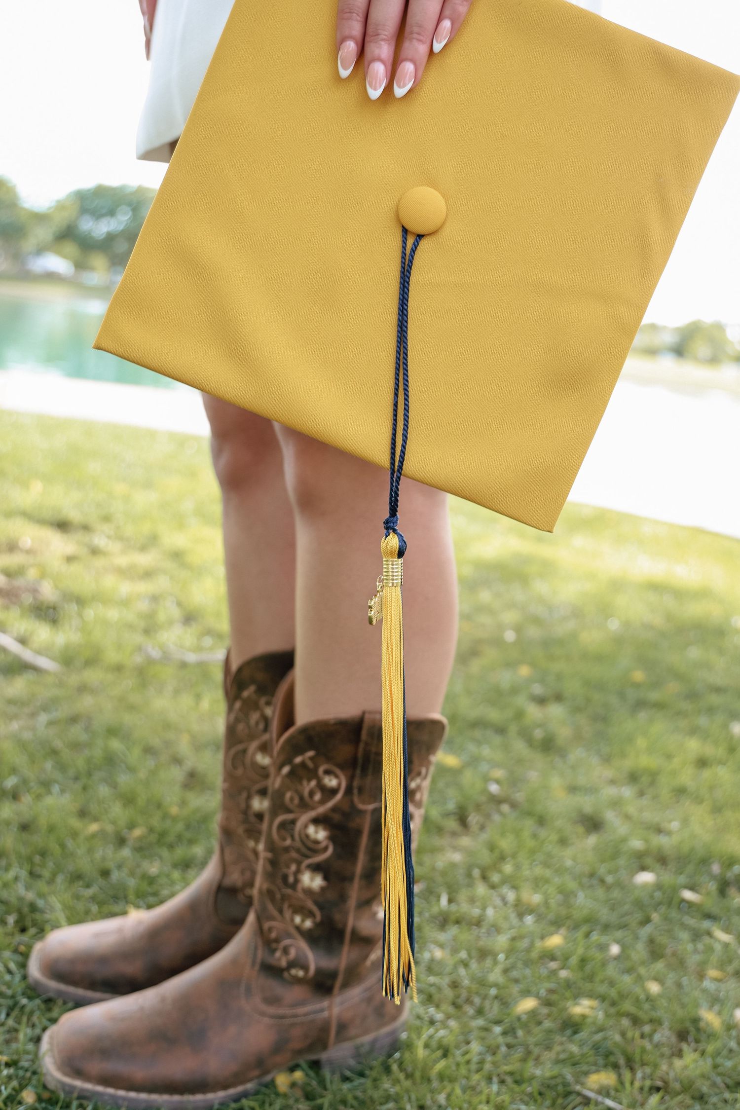 Close-up of yellow graduation cap with tassel held by graduate wearing brown cowboy boots outdoors.