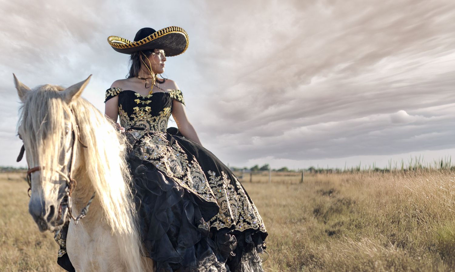 Woman in black gold embroidered gown and sombrero riding white horse in open field under cloudy sky.