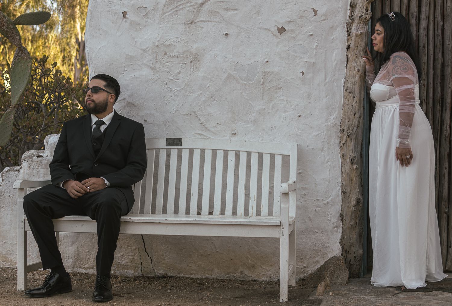 Couple sitting on white bench against rustic wall during wedding photoshoot. At Rancho Carrillo