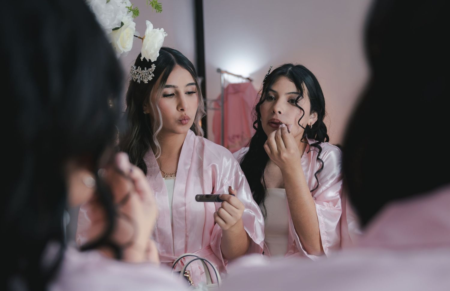 Two bridesmaids in pink satin robes apply makeup together in a mirror reflection before a wedding ceremony.