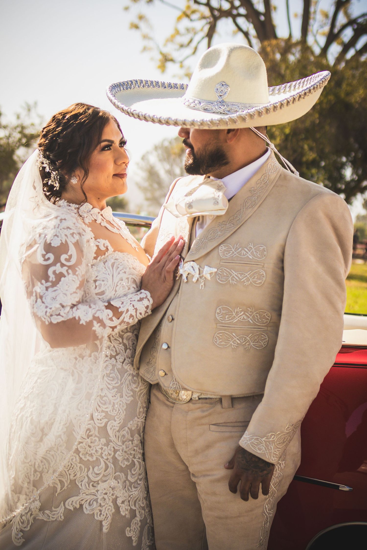 Wedding couple in Mexican-inspired attire posing with vintage red car at sunset. At San Luis Rey Parish celebrating a wedding
