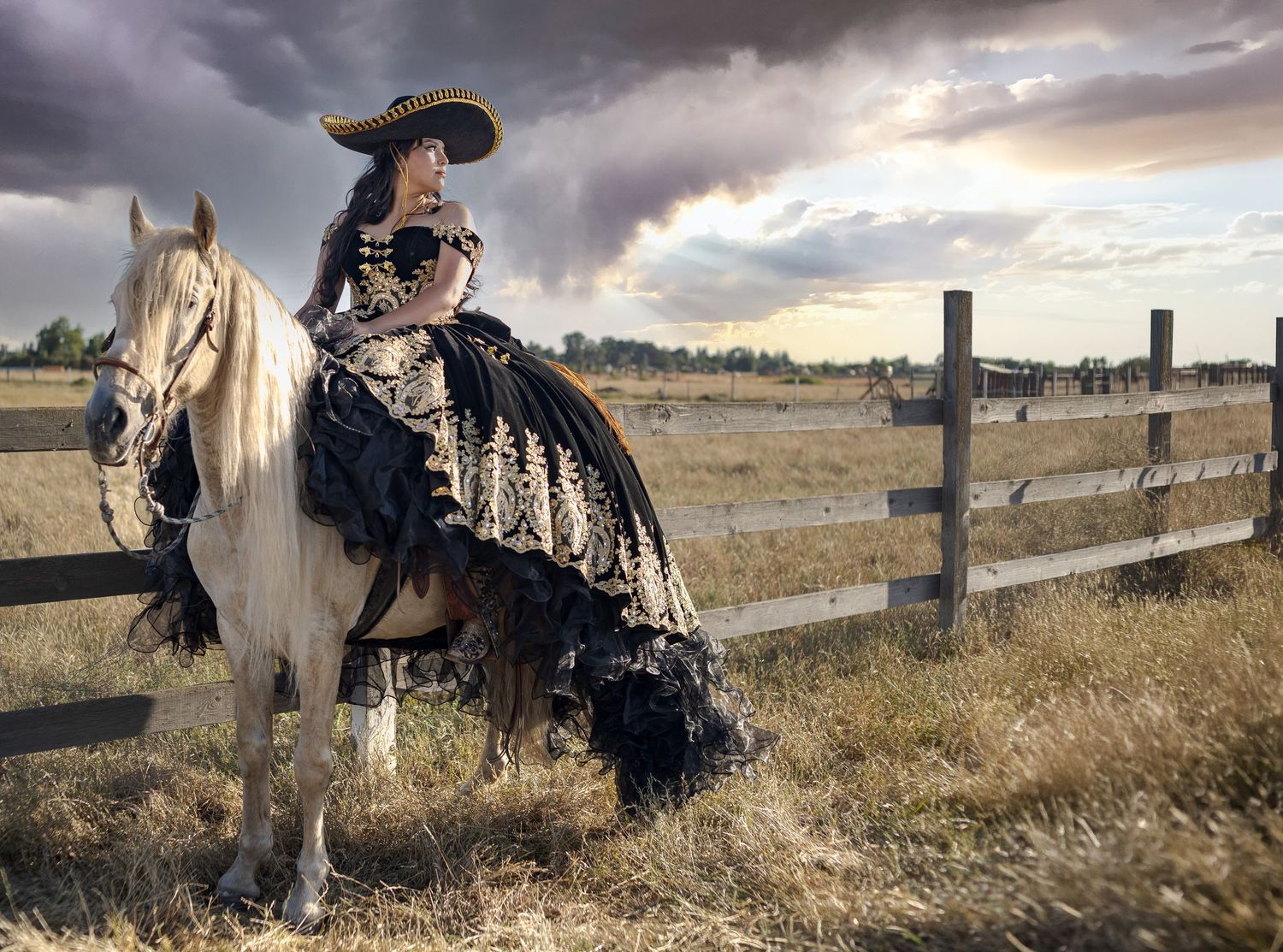 Quinceañera in Regional dress and hat riding a white horse by a wooden fence under dramatic stormy skies.
