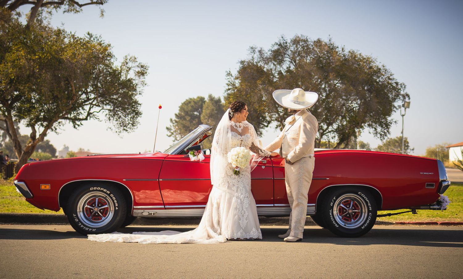 Panoramic wedding scene with vintage red car and couple against natural backdrop. At San Luis Rey Parish celebrating a wedding