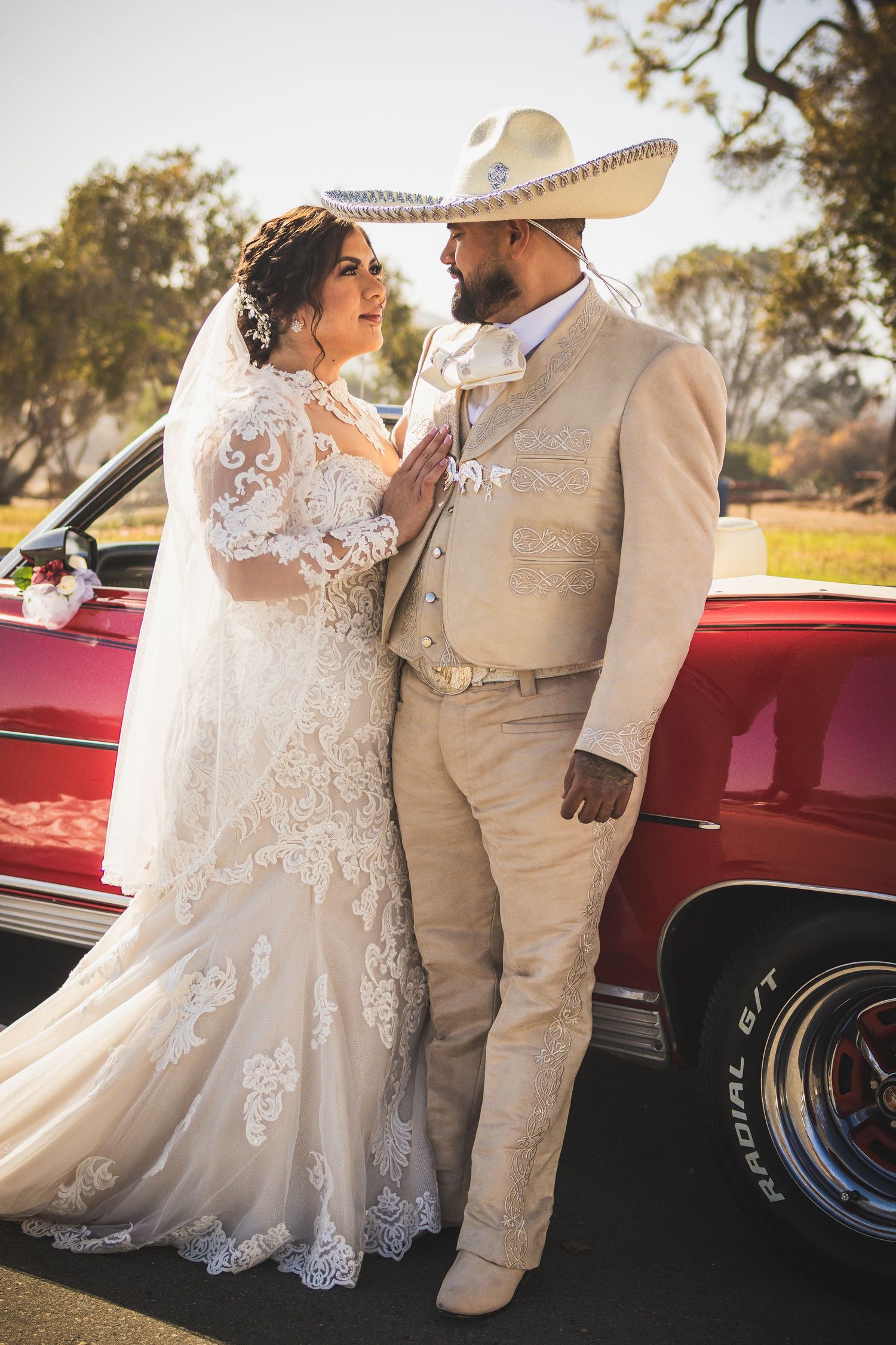 Wedding couple in elegant attire posing beside classic red car during sunset photoshoot. At San Luis Rey Parish celebrating a wedding
