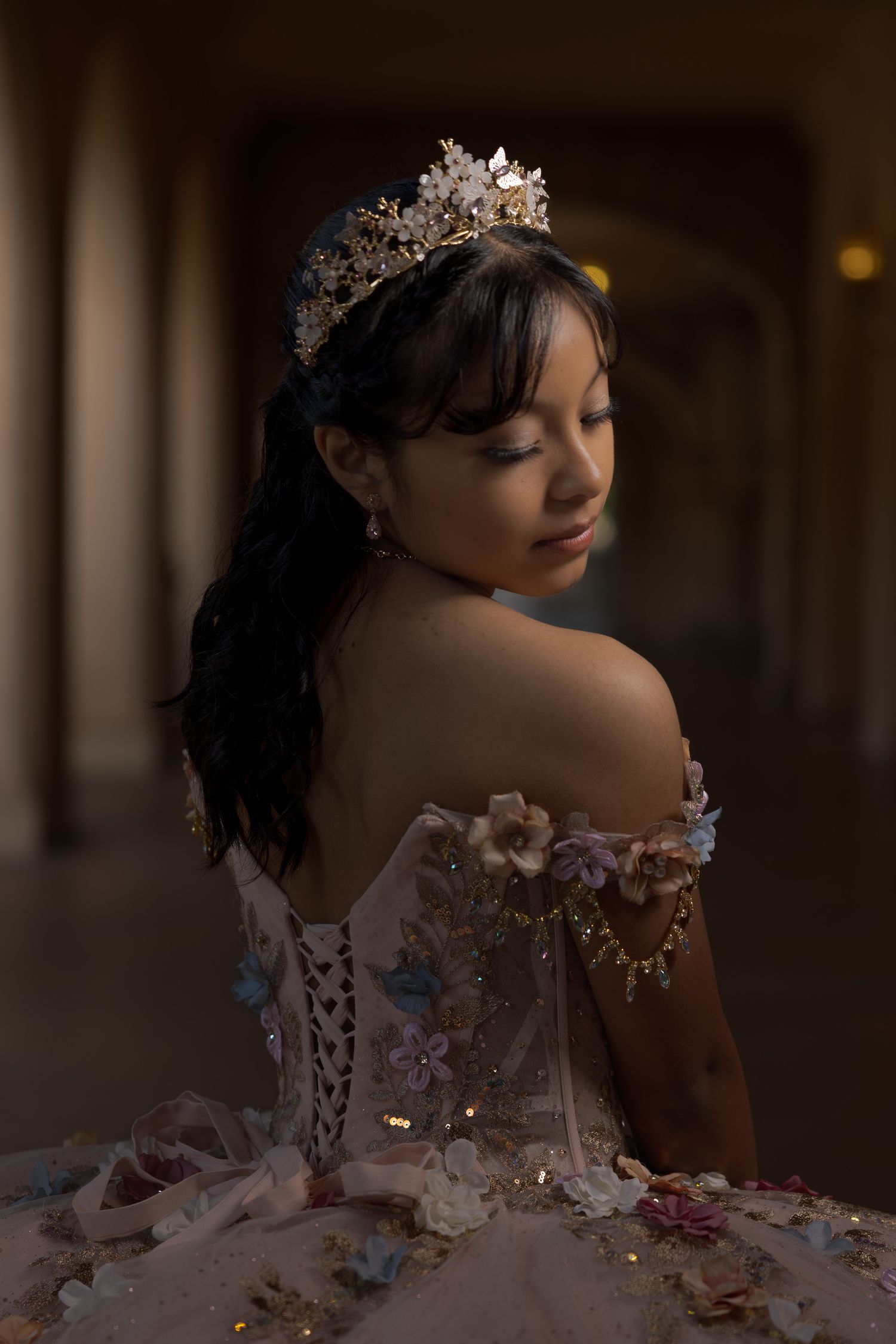 Quinceañeran in an off-shoulder floral ball gown and crown poses gracefully in a dimly lit hallway.
